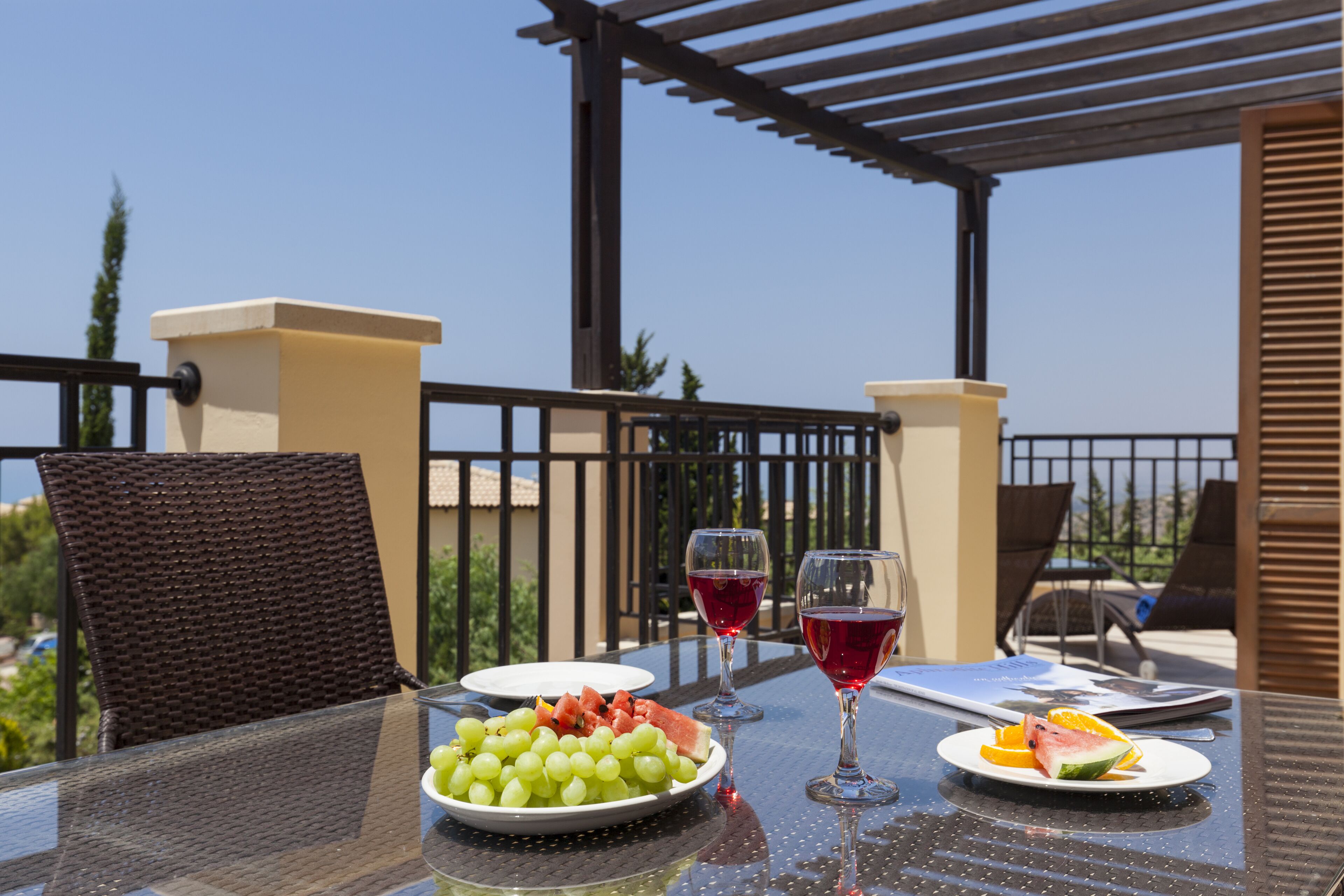 Une table à plateau en verre sur laquelle sont disposés une assiette de raisins, des tranches de pastèque et d’orange, deux verres de vin rouge, ainsi qu’un menu. La table se trouve sur un balcon agrémenté de chaises en osier et d’une pergola.