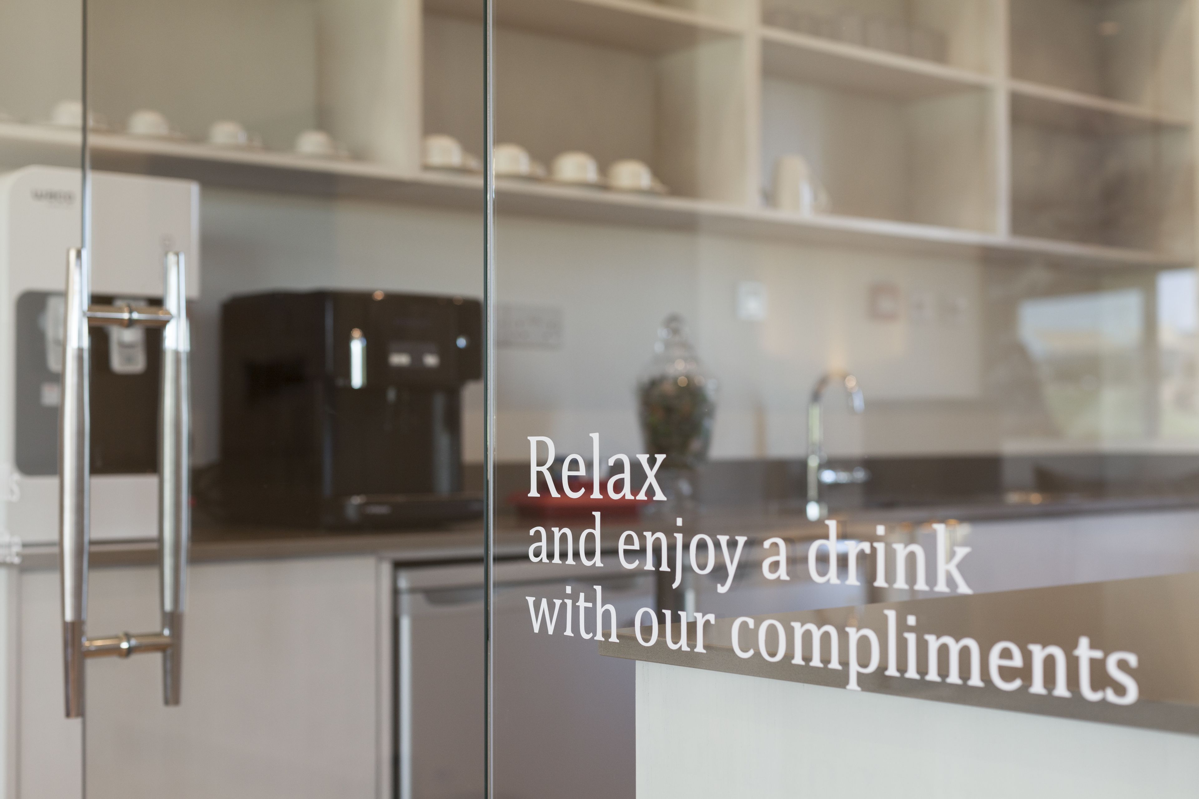 A modern kitchen with a coffee machine and a glass door with a message about enjoying a drink with compliments.
