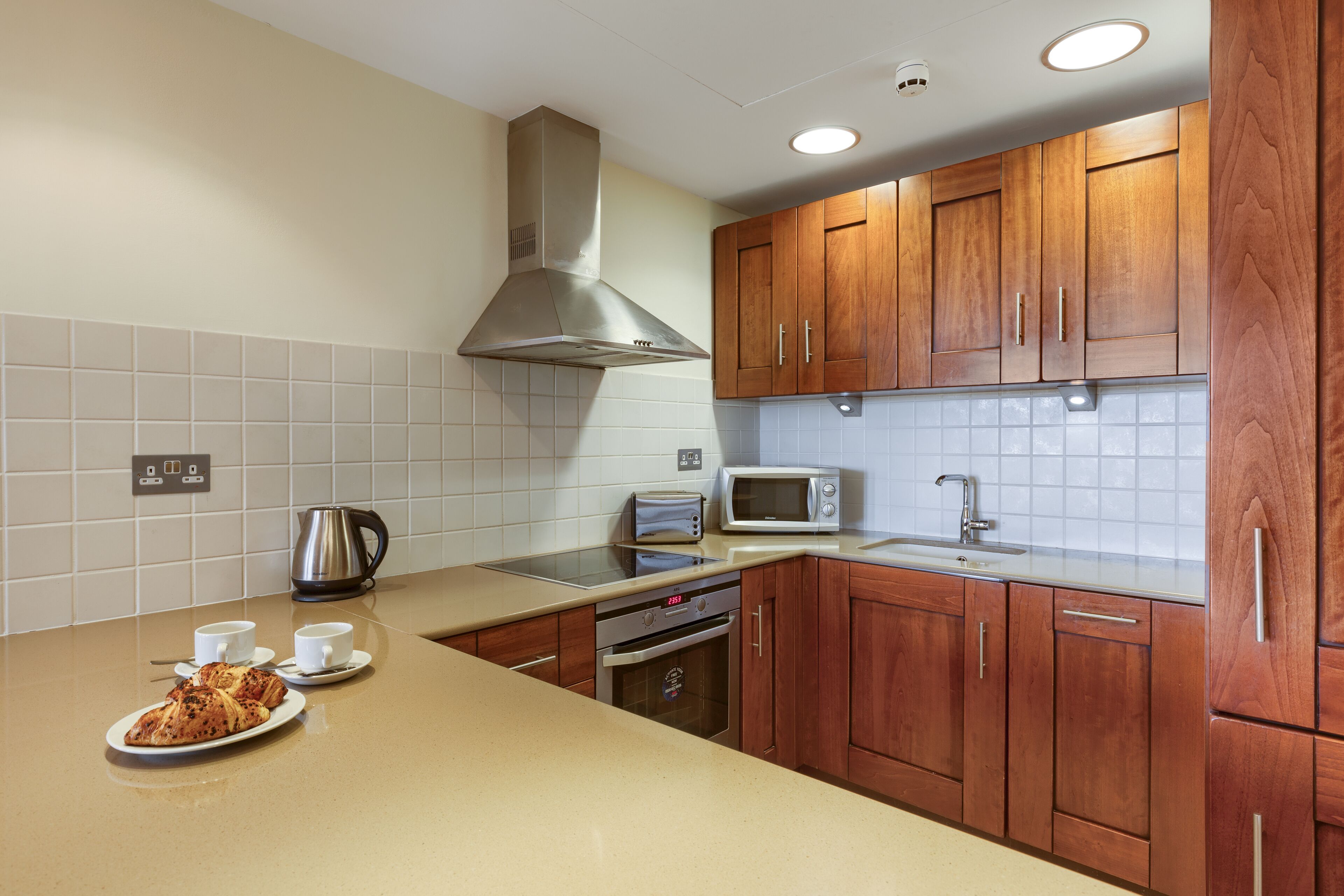 A kitchen with wooden cabinets, a stainless steel range hood, a microwave, a kettle, and a plate with croissants and cups.