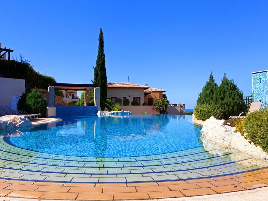 A swimming pool with a tiled edge, a pergola, and a view of a building with a tiled roof.