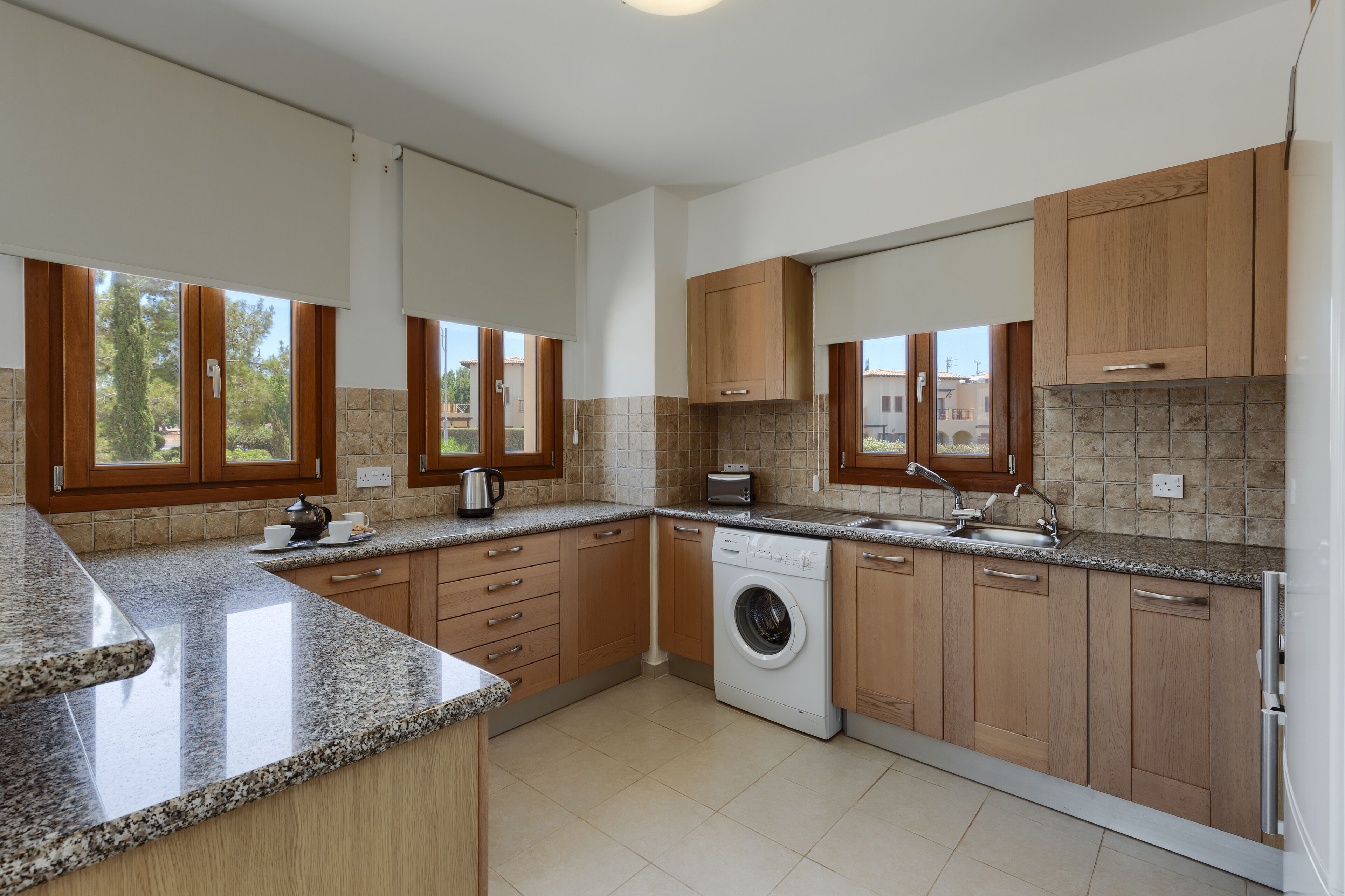 Una cocina moderna con encimeras de granito, armarios de madera, lavadora integrada y un amplio ventanal que proporciona luz natural.