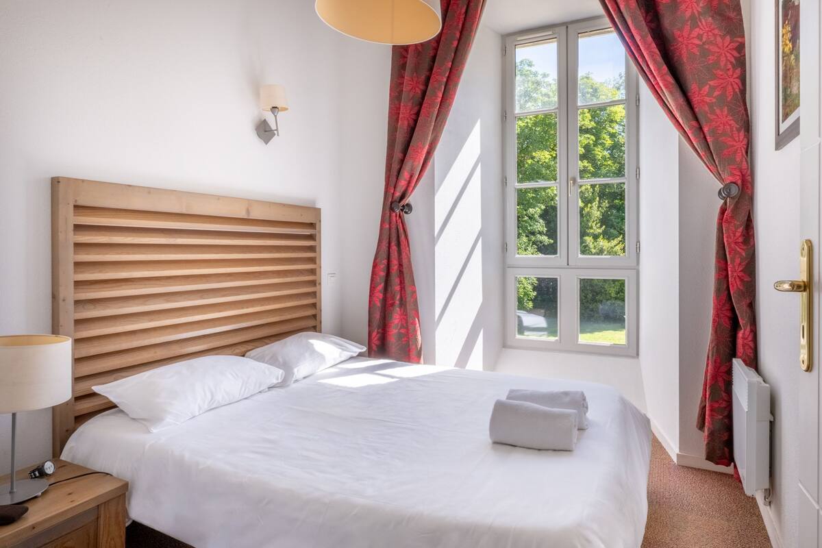 Un dormitorio con cabecera de madera, cama con ropa blanca, mesita de noche con lámpara y cortinas rojas.