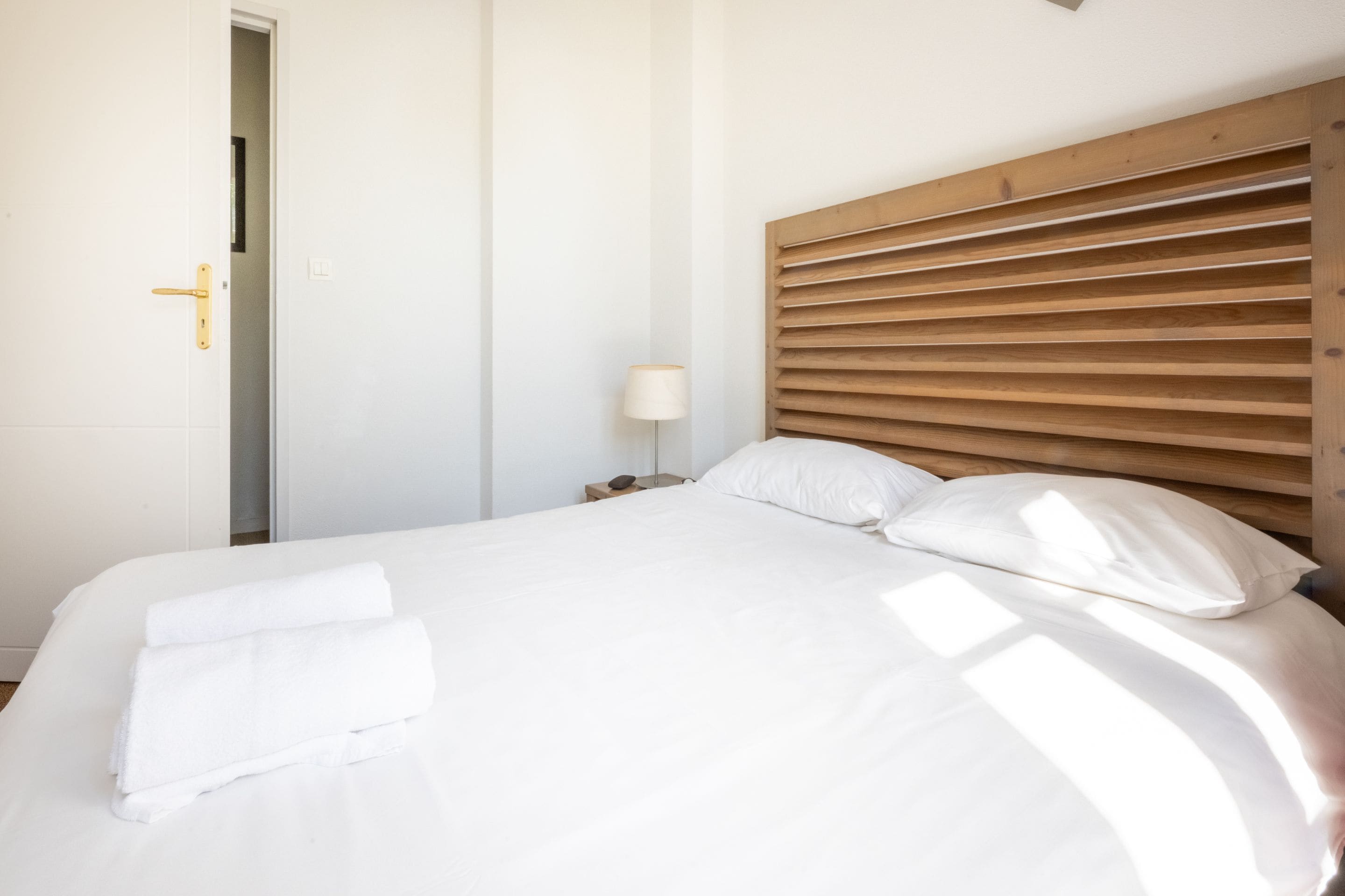 Un lit bien fait, avec du linge de lit blanc, une tête de lit en bois et une lampe de chevet.