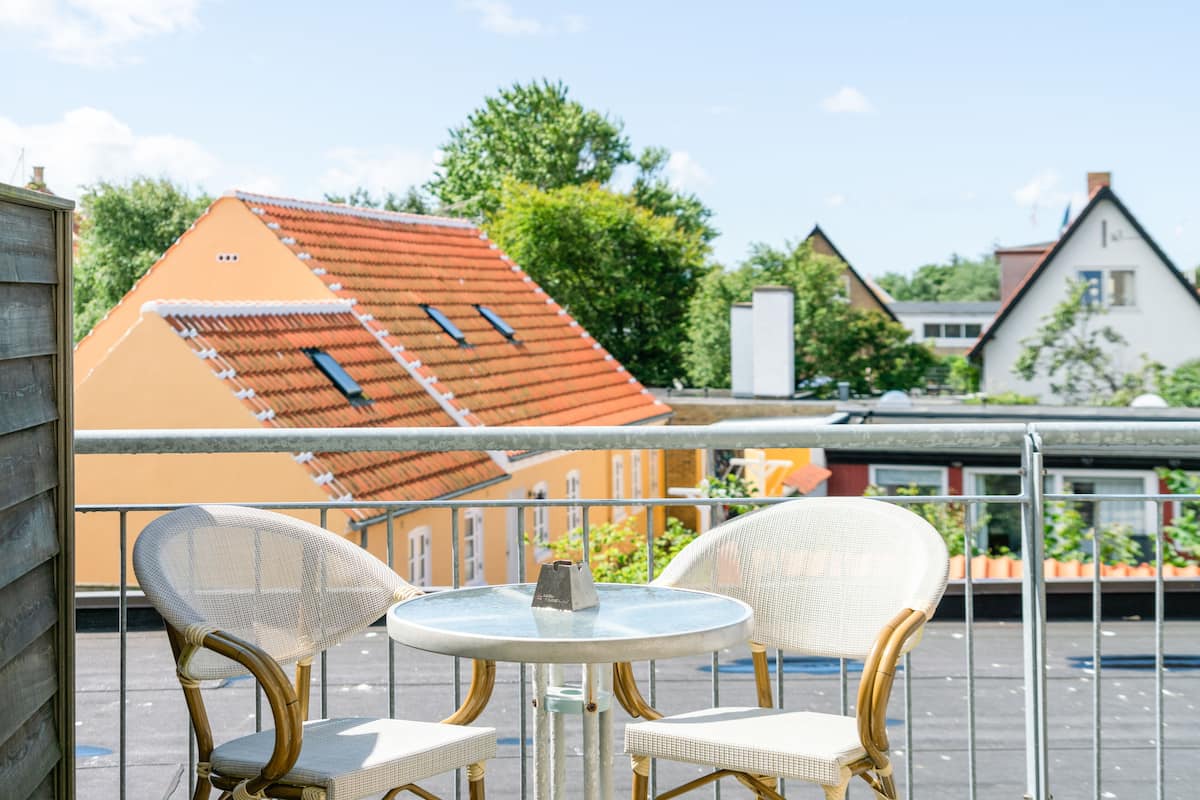 Een balkon met twee rieten stoelen en een kleine ronde tafel, met uitzicht op een woonwijk met huizen en bomen.