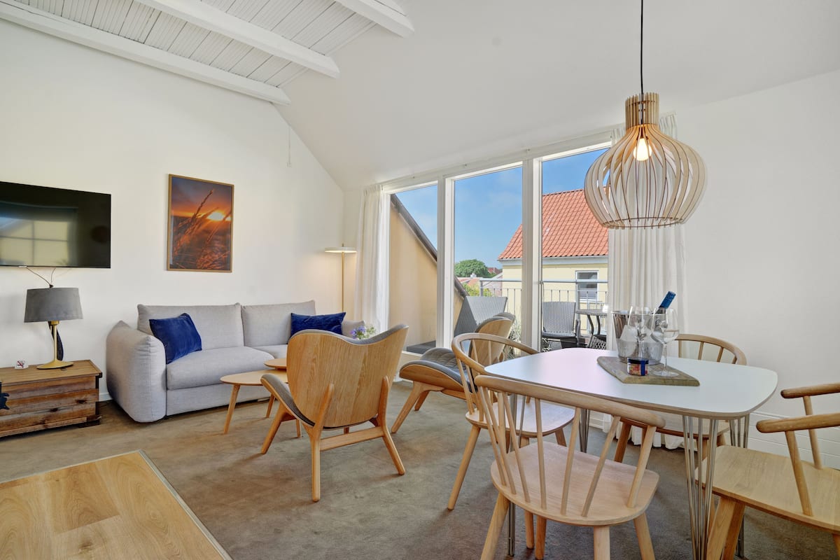 Een moderne woonkamer met een bank, stoelen, een eettafel en een groot raam met uitzicht op een gebouw.