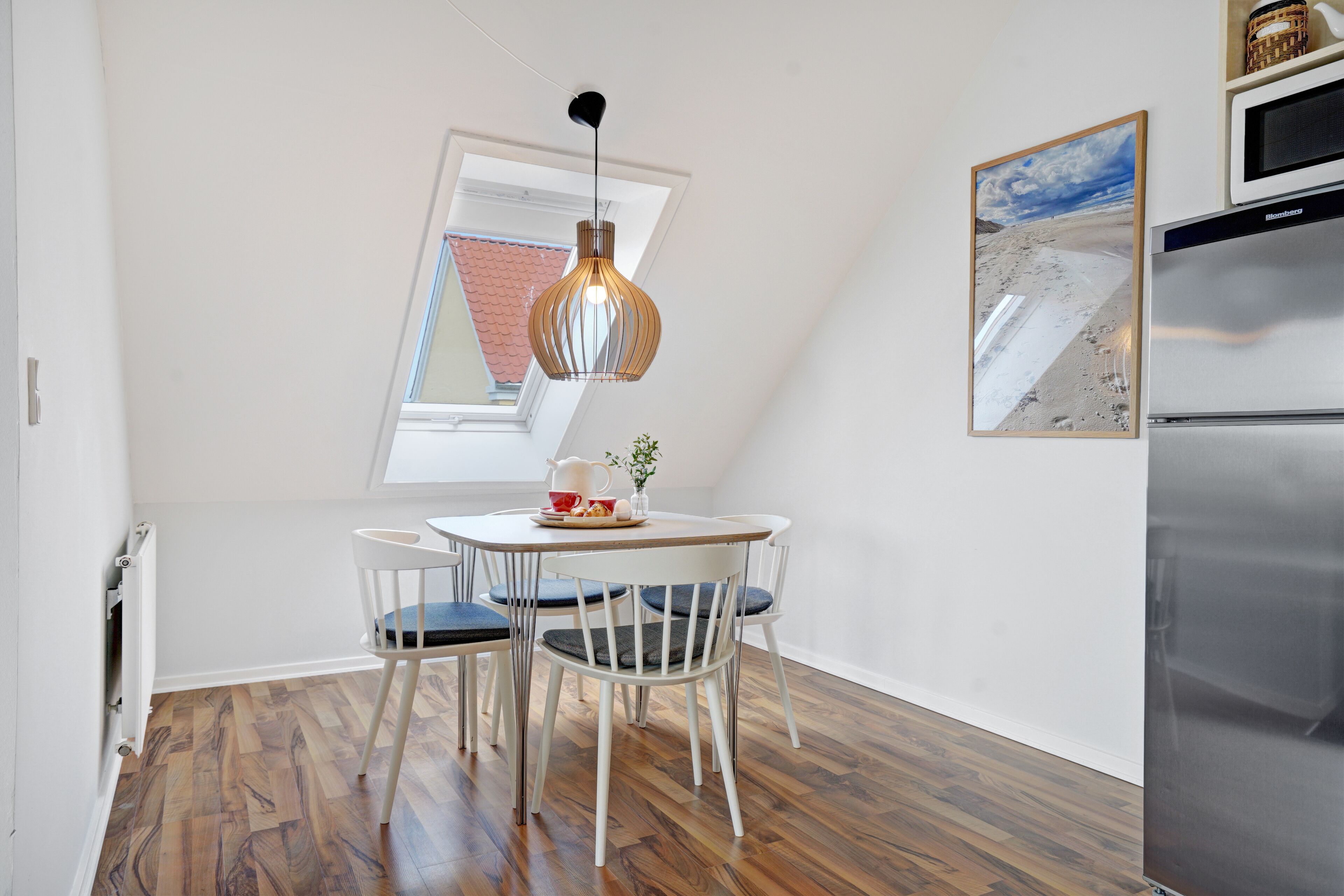 A small kitchen with a round table, white chairs, a hanging light fixture, a refrigerator, and a framed picture on the wall.