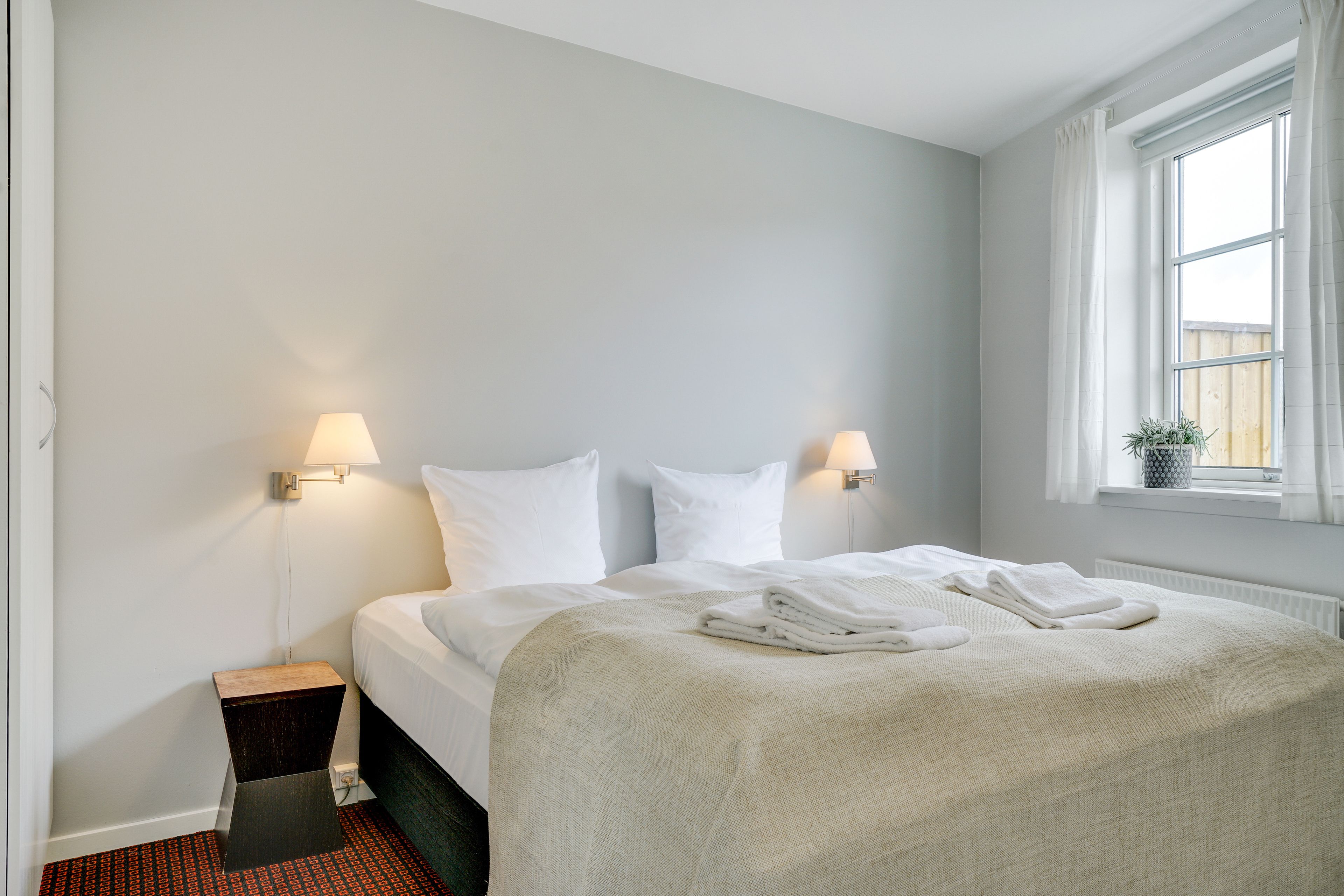 A neatly made bed with white linens, a bedside table with a lamp, and a window with curtains.
