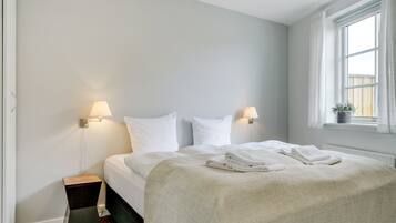 A neatly made bed with white linens, a bedside table with a lamp, and a window with curtains.