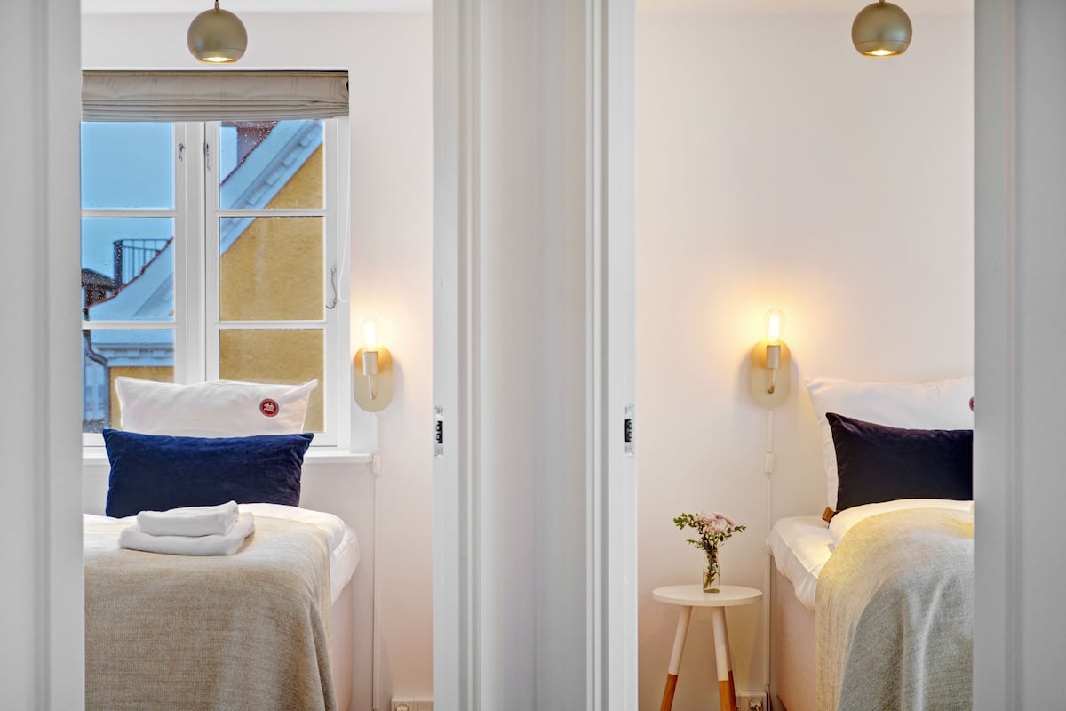 Een hotelkamer met twee bedden, elk met een ander kussendesign. Er zijn wandlampen en een klein tafeltje met een vaas.