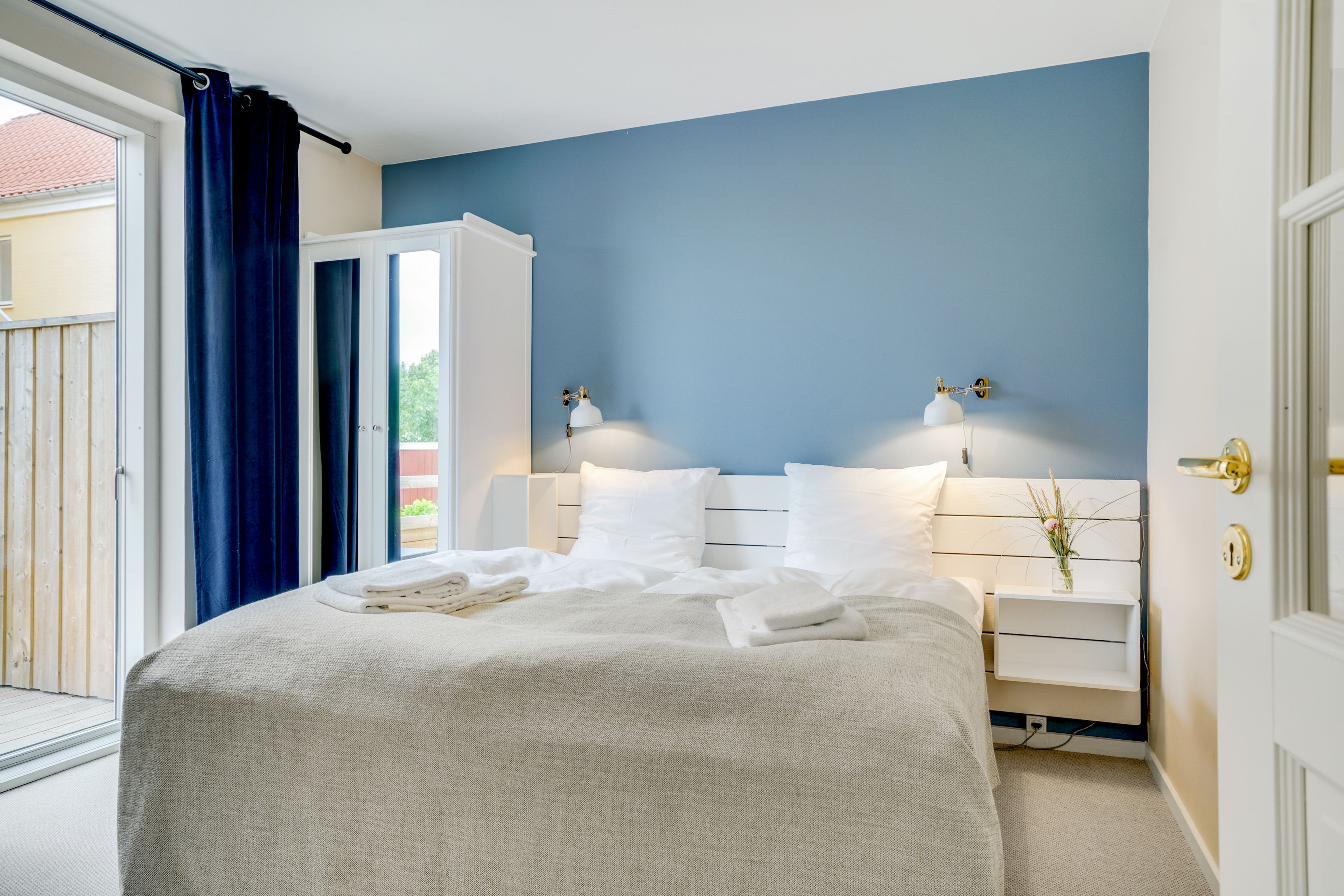 A neatly made bed with a white comforter and pillows, a bedside table with a lamp, and a view of the outdoors through a sliding door.