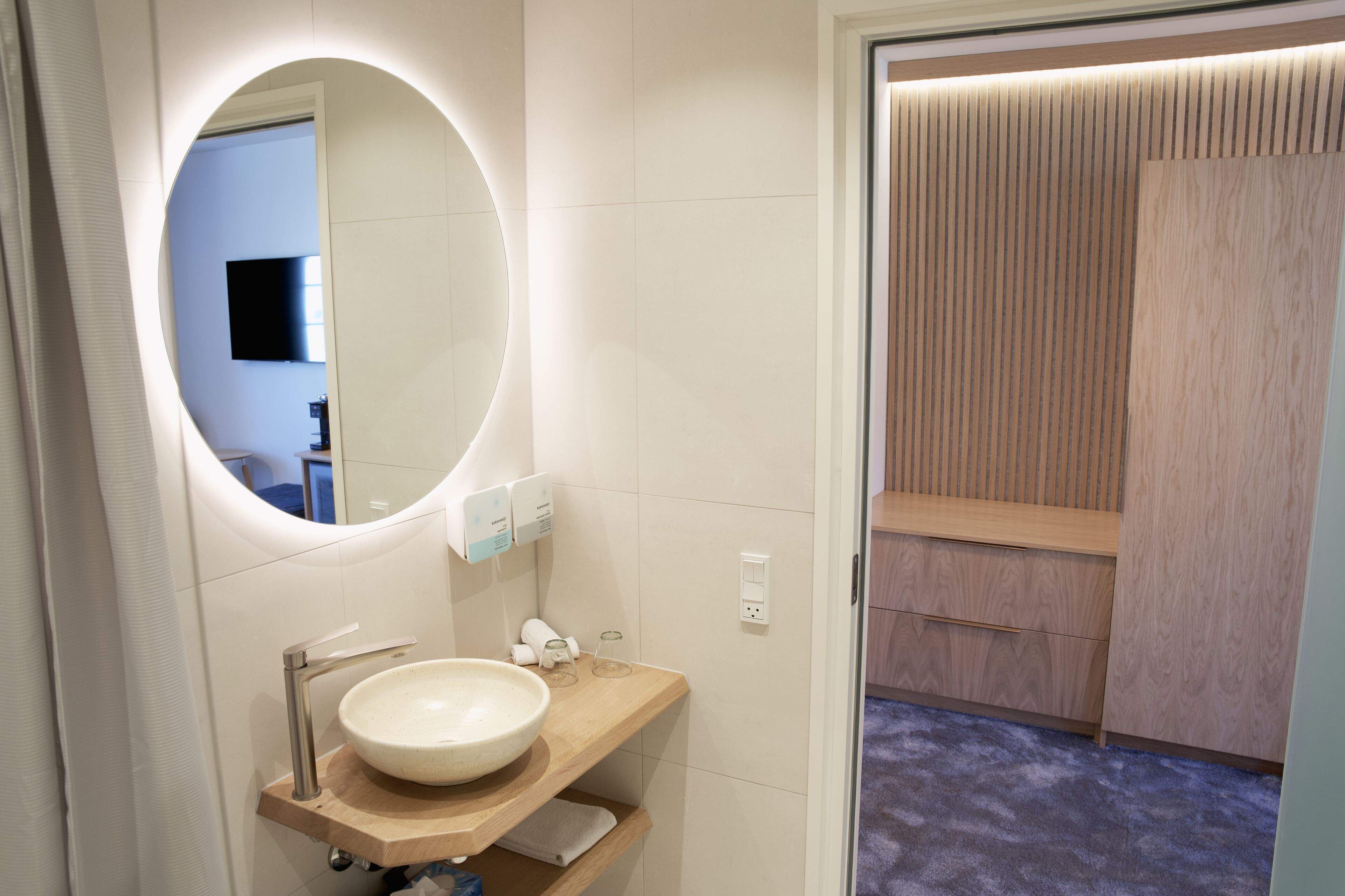 Un baño moderno con un espejo redondo, un mueble de tocador de madera, un lavabo y una zona de ducha con paneles de madera.