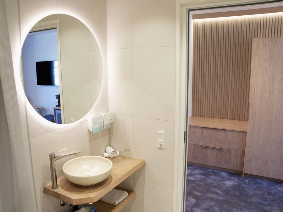 Un baño moderno con un espejo redondo, un mueble de tocador de madera, un lavabo y una zona de ducha con paneles de madera.