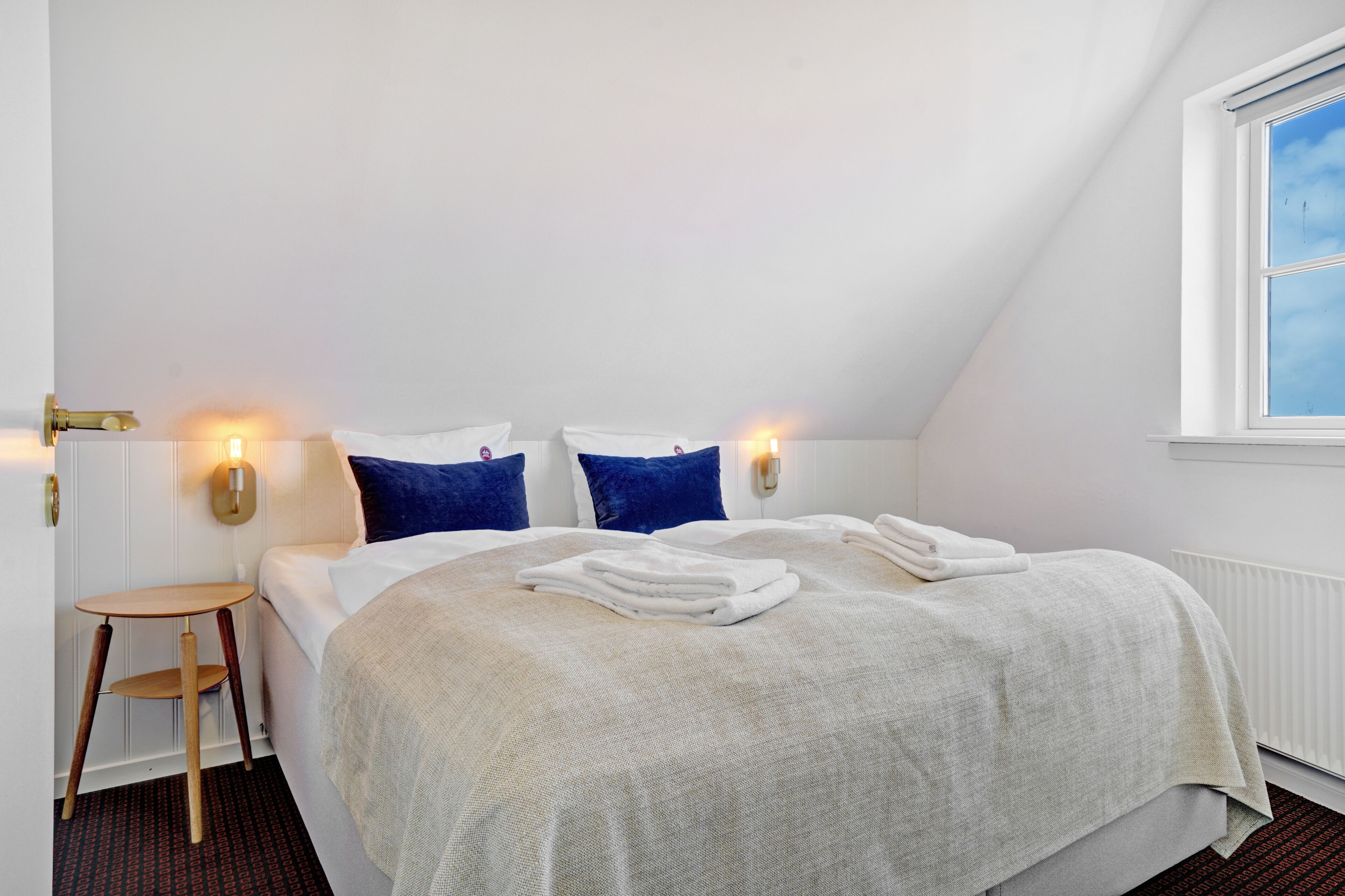 Una cama bien hecha con ropa de cama blanca y azul, una mesita de madera y una lámpara de pared.