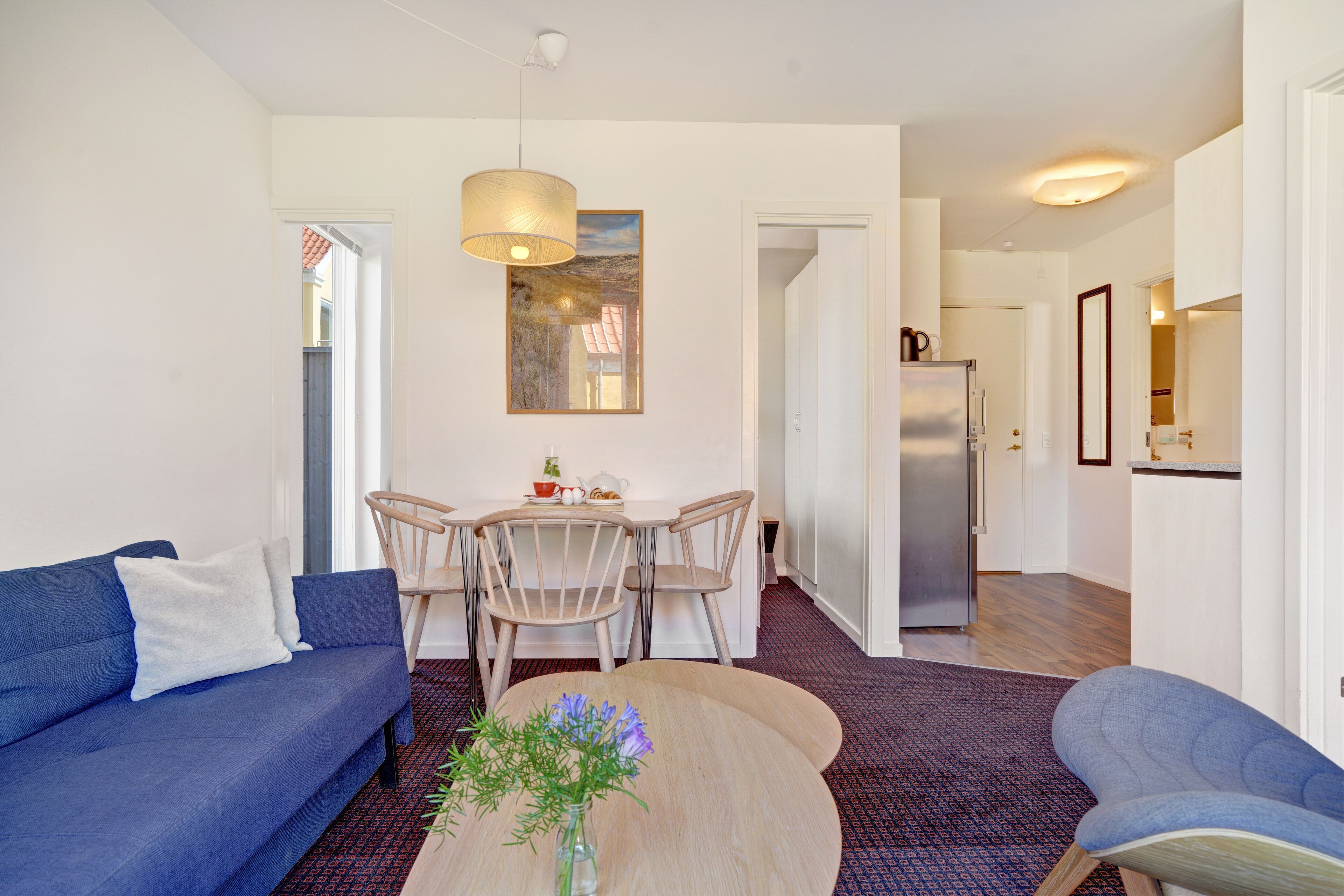 Ein modernes Wohnzimmer mit einem blauen Sofa, einem Couchtisch mit Blumen, einem Essbereich mit Tisch und Stühlen sowie einer Küche mit Kühlschrank.