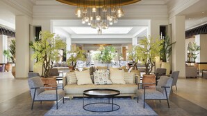 A spacious lobby with a central seating area featuring a sofa and armchairs, surrounded by potted plants and a modern chandelier.