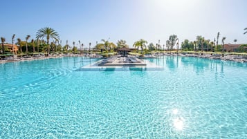 Una piscina amplia y cristalina con sillas de descanso y sombrillas, rodeada de palmeras y un cielo despejado.