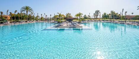 A large, clear swimming pool with lounge chairs and umbrellas, surrounded by palm trees and a clear sky.