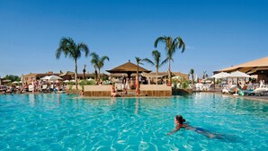 A large outdoor swimming pool with clear blue water, surrounded by palm trees and umbrellas.
