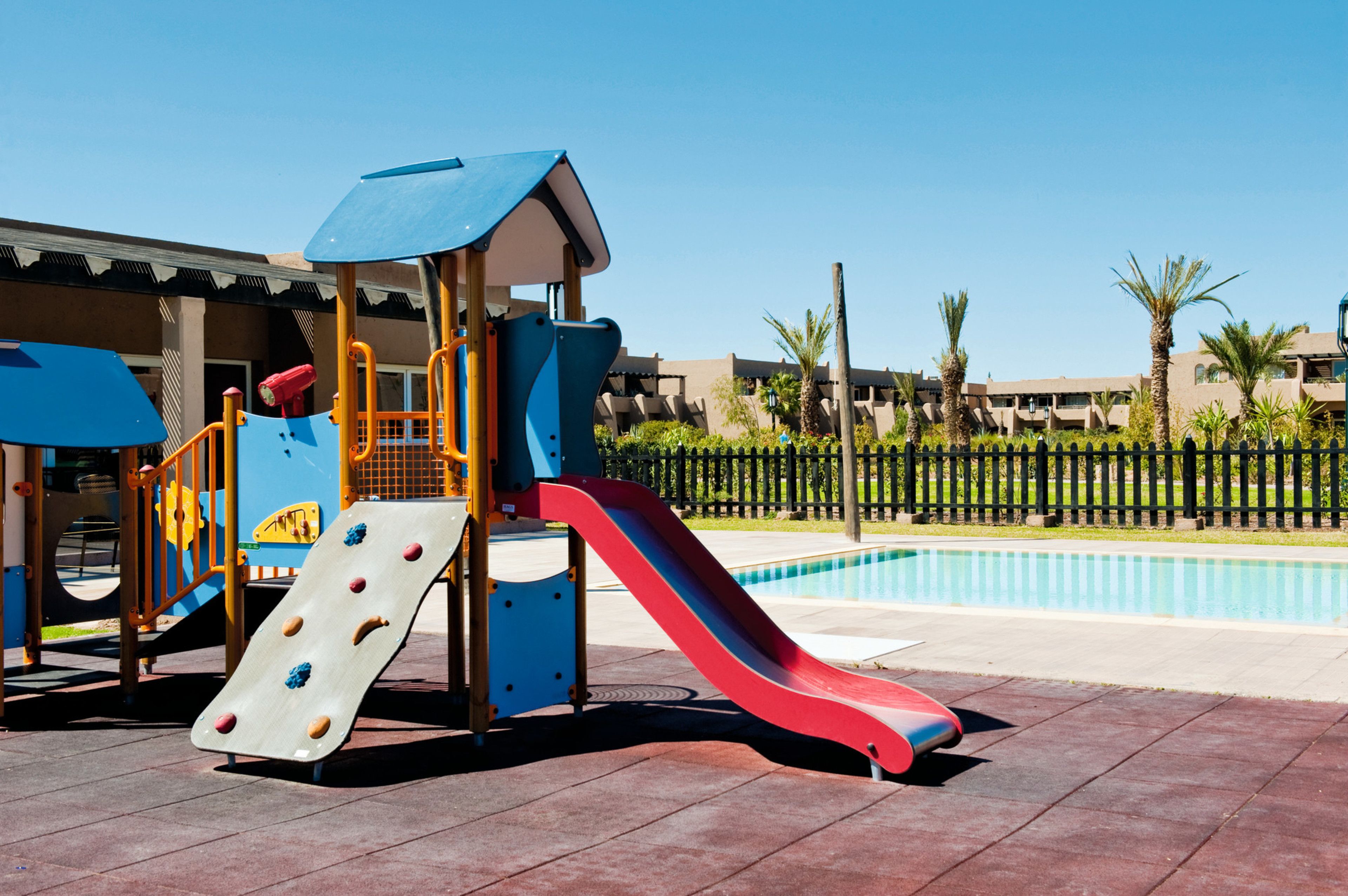 Un terrain de jeux avec un toboggan et une zone d’escalade, une piscine et un bâtiment doté de balcons en arrière-plan.