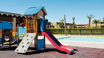 A playground with a slide and climbing area, a pool, and a building with balconies in the background.