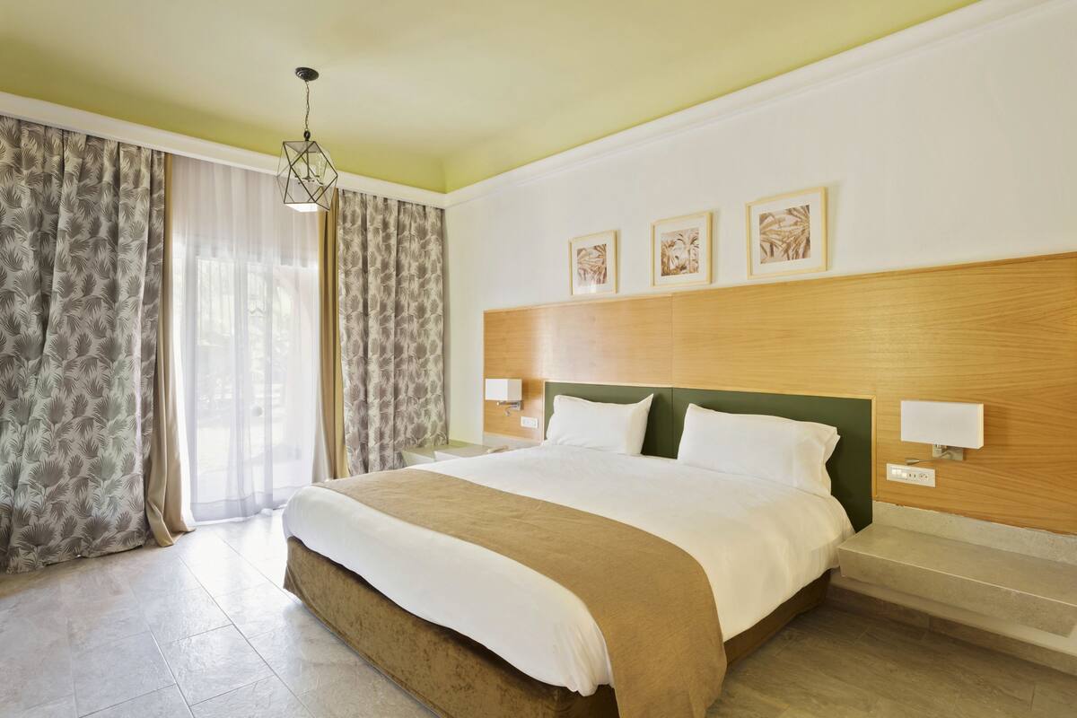 A hotel room with a large bed, a wooden headboard, a nightstand, and a window with sheer curtains.