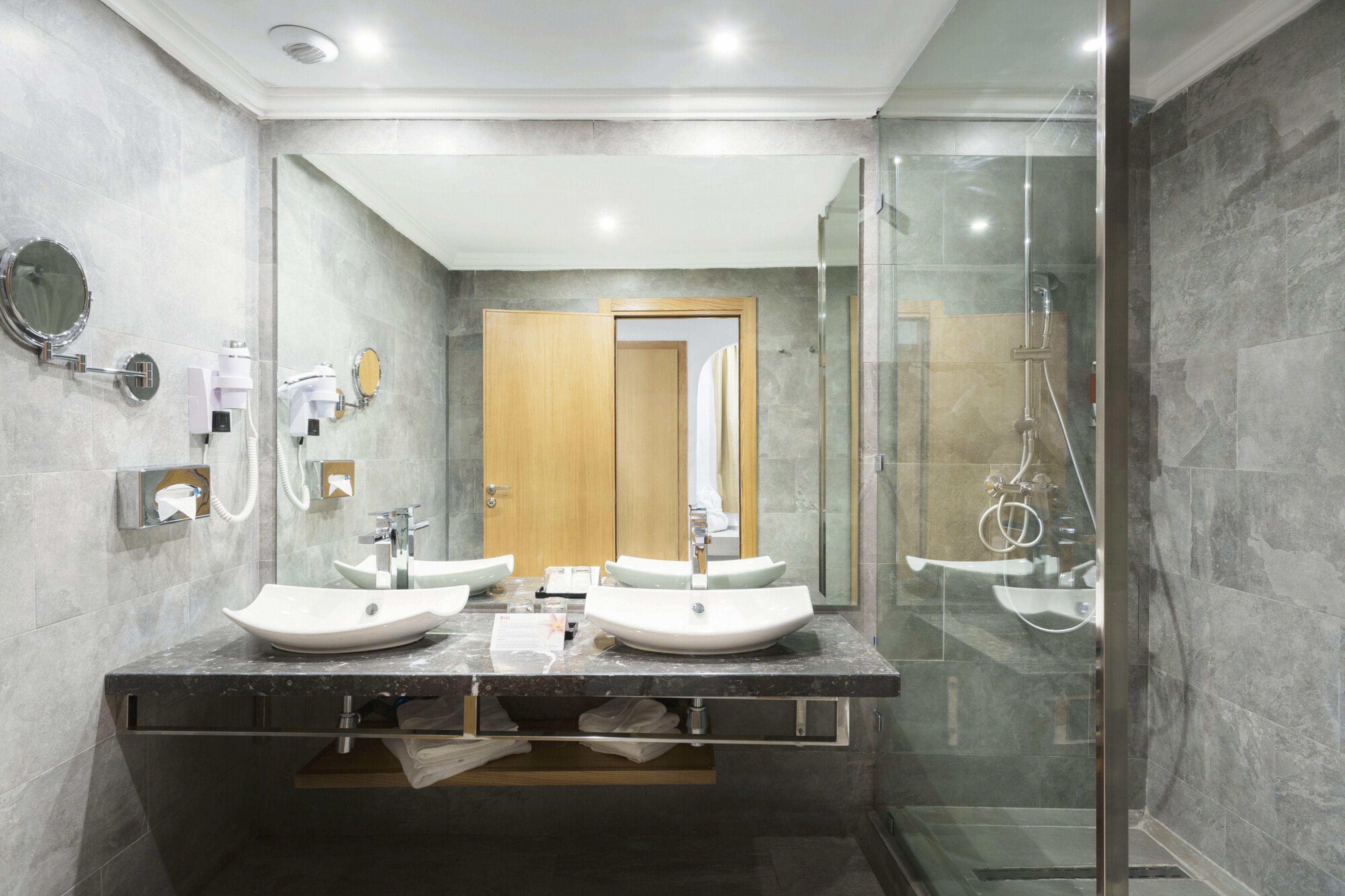 A modern bathroom with two sinks, a glass shower enclosure, and a wooden door in the background.