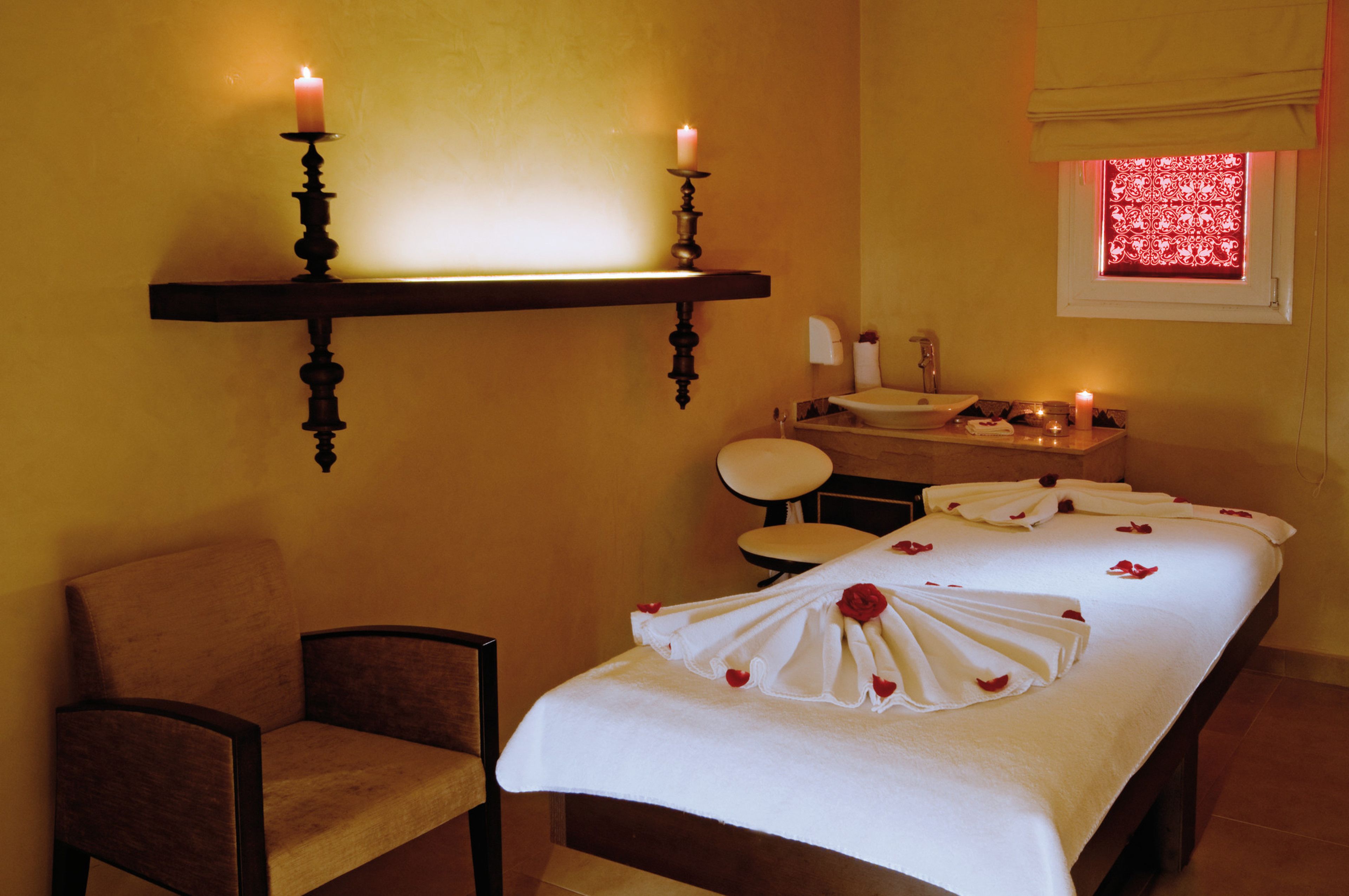 A spa room with a massage table, candles, a chair, and a window with a red pattern.