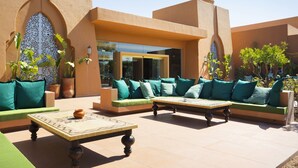 An outdoor seating area with green sofas and a wooden table, surrounded by potted plants and architectural arches.