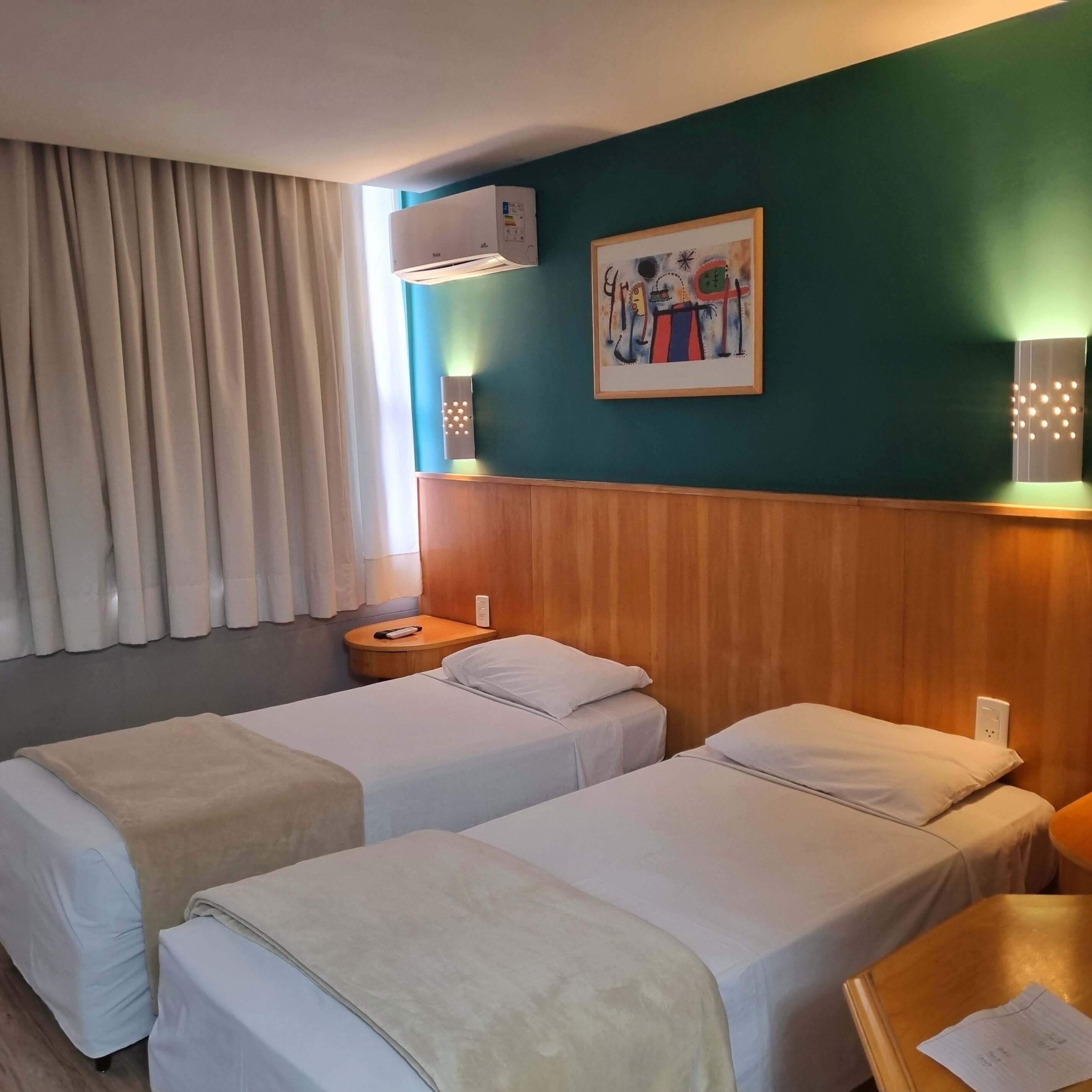 Camera d'albergo con due letti, una testiera in legno, un quadro appeso e una finestra con tende.