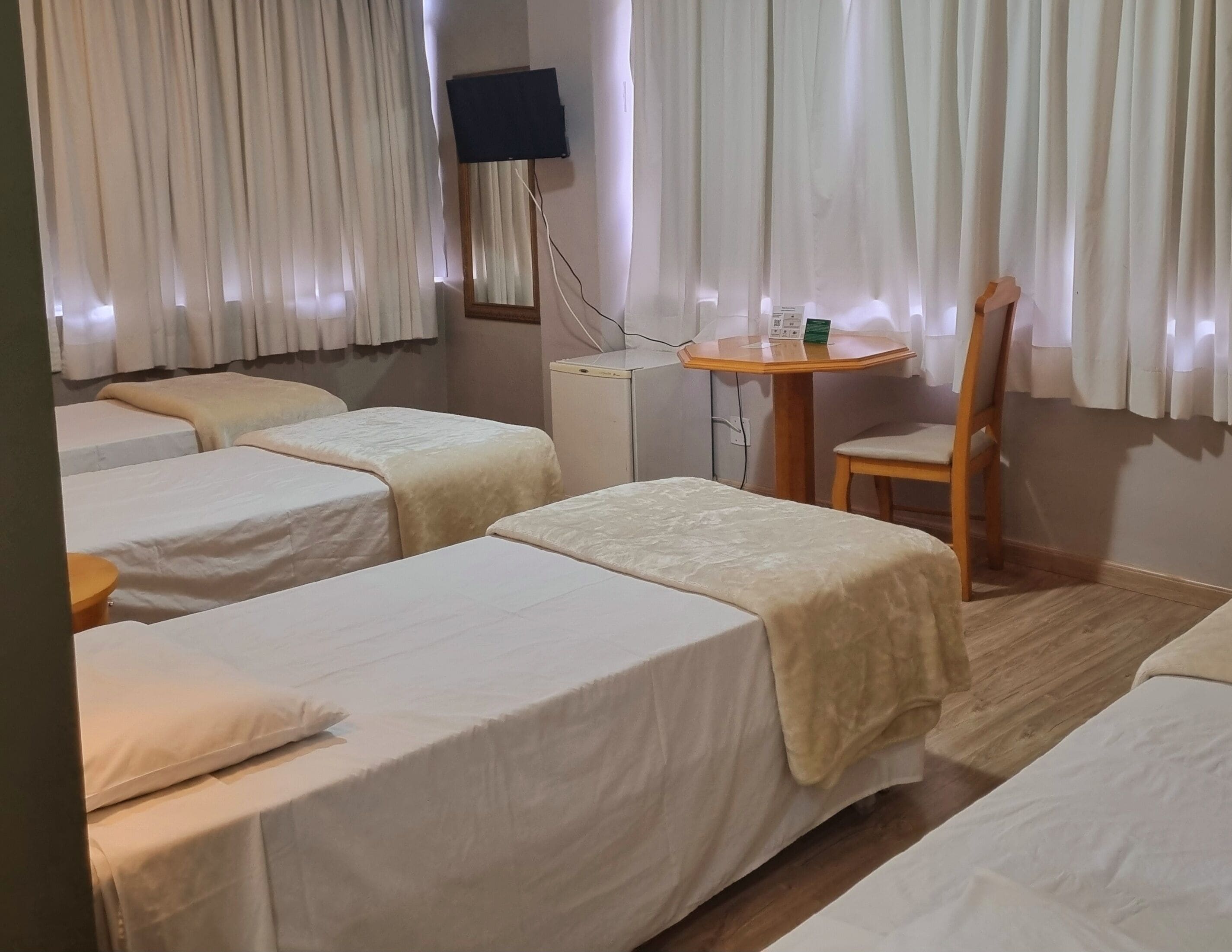 Une chambre d’hôtel avec trois lits simples, une petite table avec une chaise, un téléviseur fixé au mur et une fenêtre avec des rideaux.