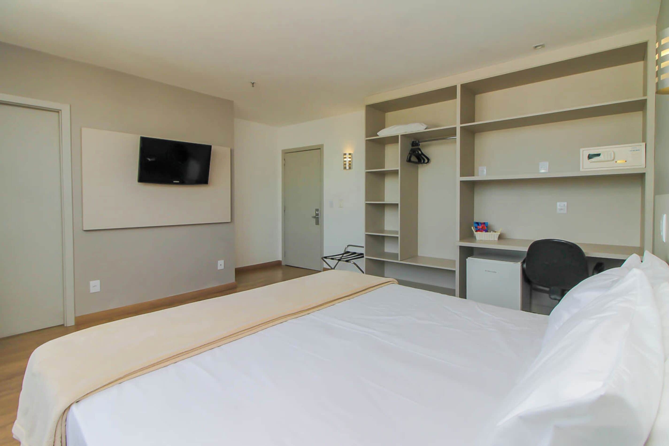 Habitación de hotel con cama, escritorio, silla, televisor y estanterías empotradas.