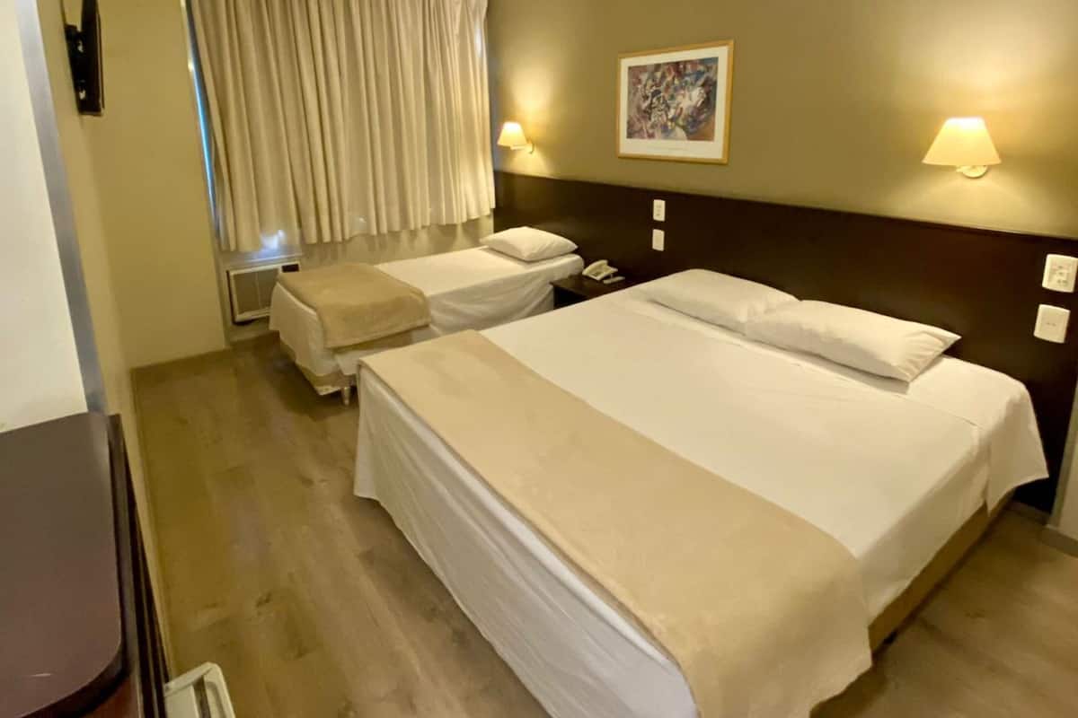 Une chambre d’hôtel avec un grand lit, un lit plus petit, un bureau et une chaise. Il y a deux lampes fixées au mur et un tableau accroché au mur.