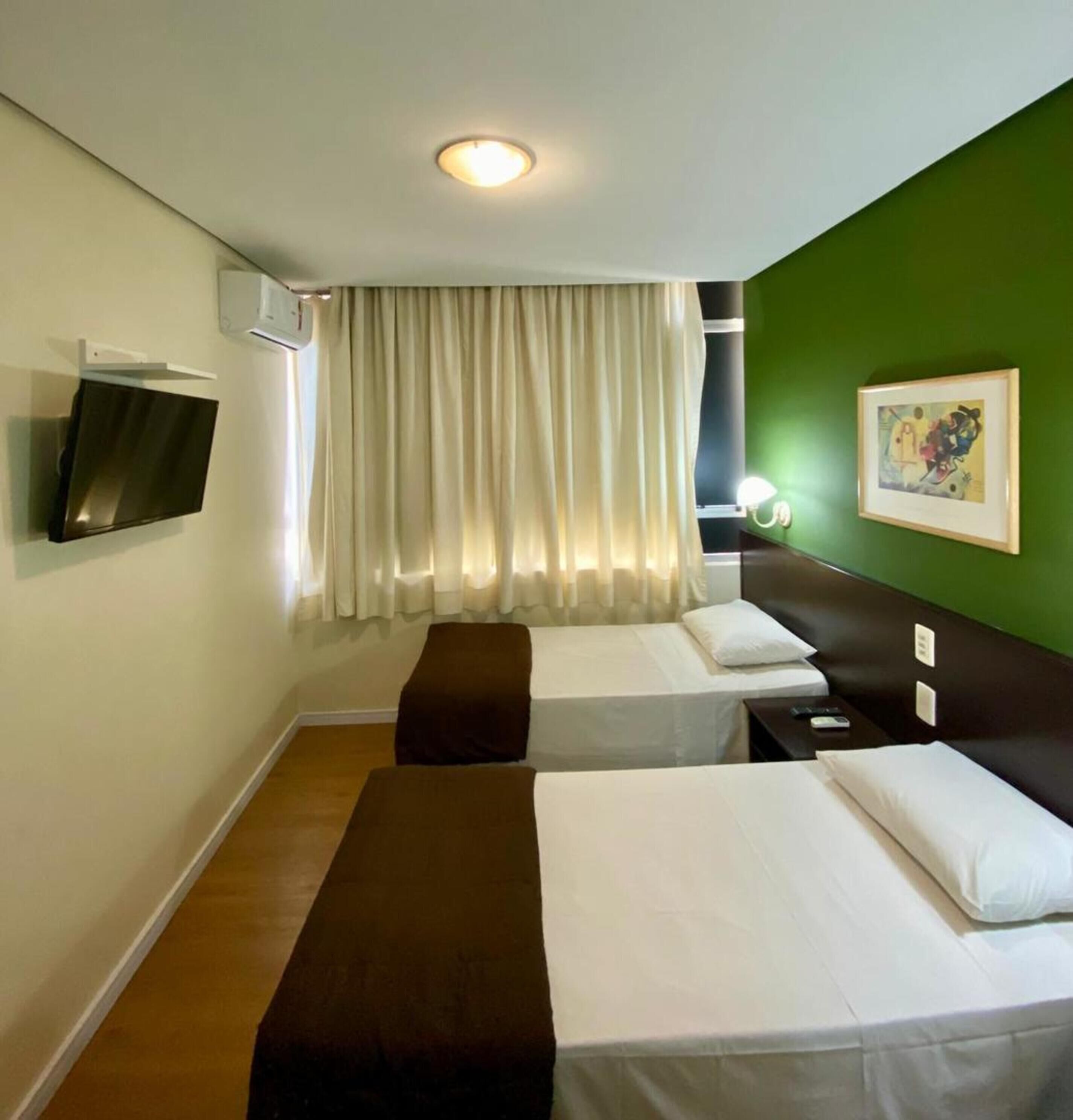 Une chambre d’hôtel avec deux lits, un téléviseur à écran plat fixé au mur, une fenêtre avec des rideaux et un mur d’accent de couleur verte.