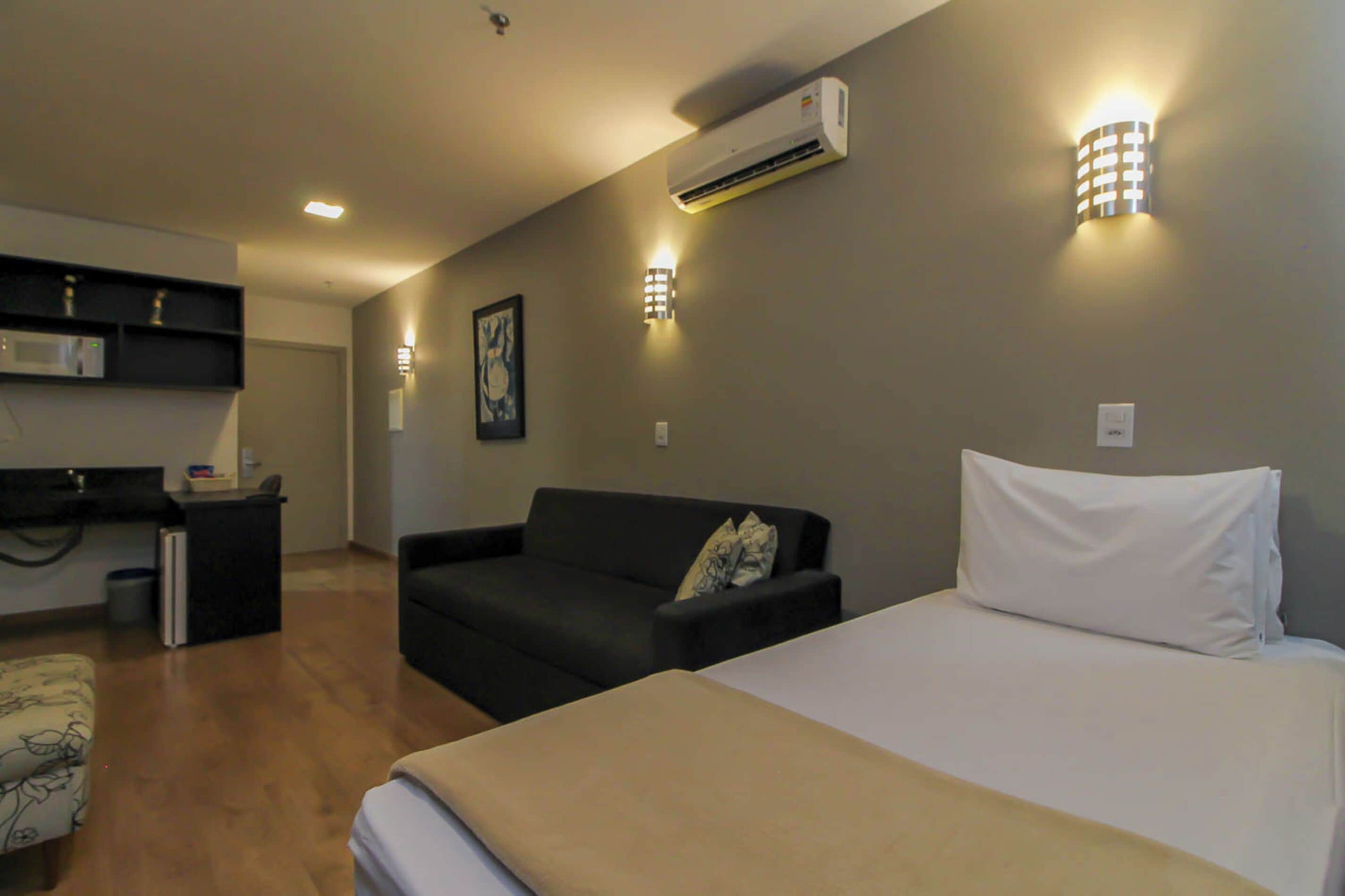 A hotel room with a bed, a sofa, a small table, and an air conditioning unit on the wall.