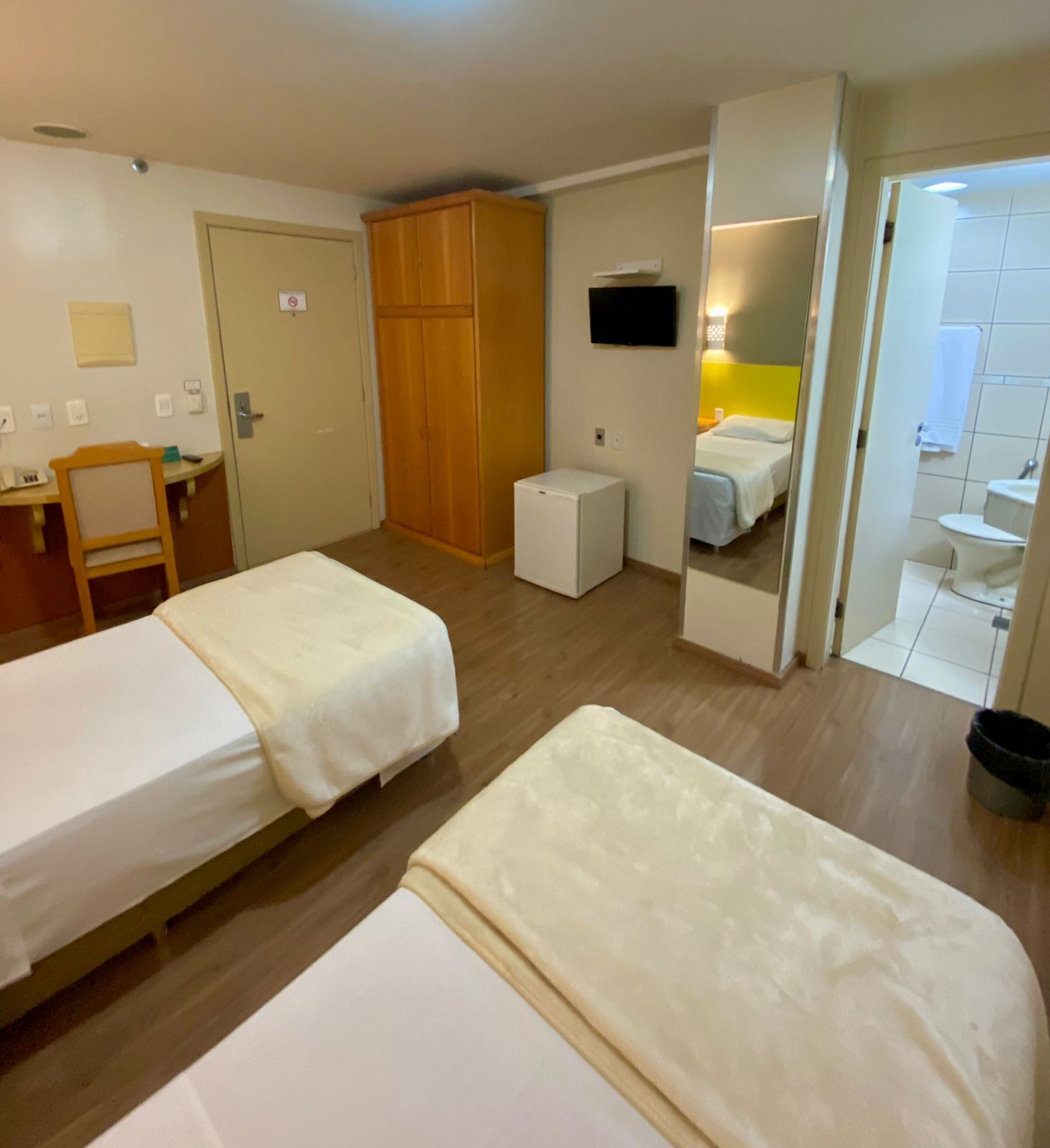 Habitación de hotel con dos camas individuales, un armario de madera, un escritorio pequeño con una silla, un televisor montado en la pared y un baño visible a través de una puerta abierta.