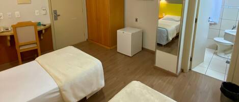 Habitación de hotel con dos camas individuales, un armario de madera, un escritorio pequeño con una silla, un televisor montado en la pared y un baño visible a través de una puerta abierta.