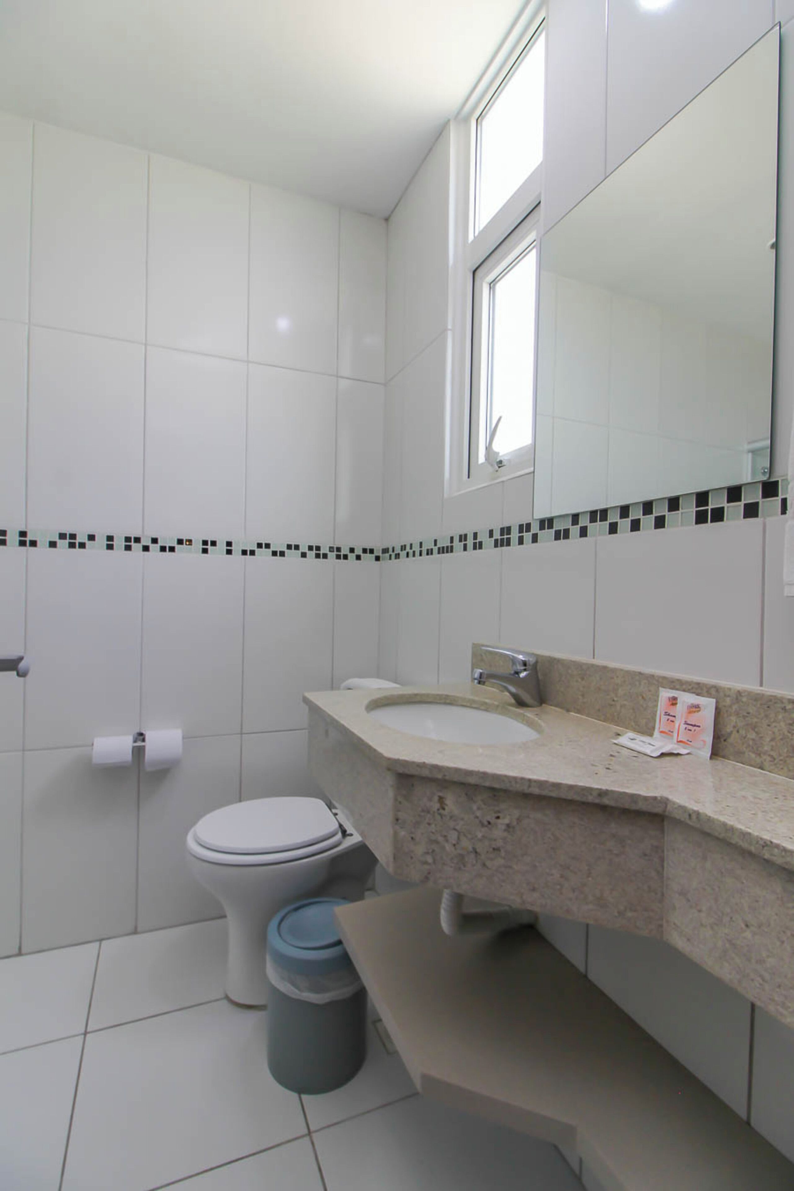 A bathroom with white tiles, a toilet, a sink, a mirror, and a trash bin.
