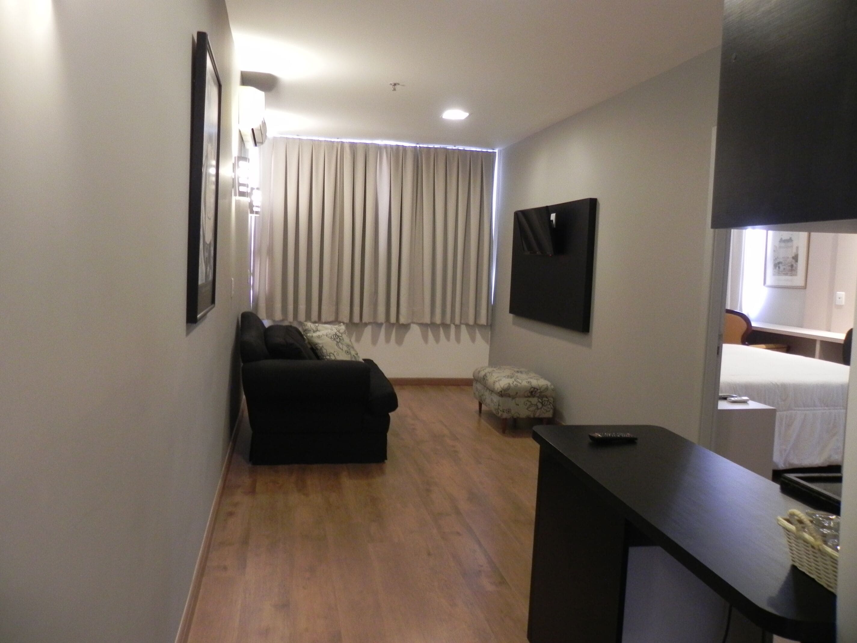 A compact living space with a sofa, a small ottoman, a flat-screen TV mounted on the wall, and a desk with a basket.