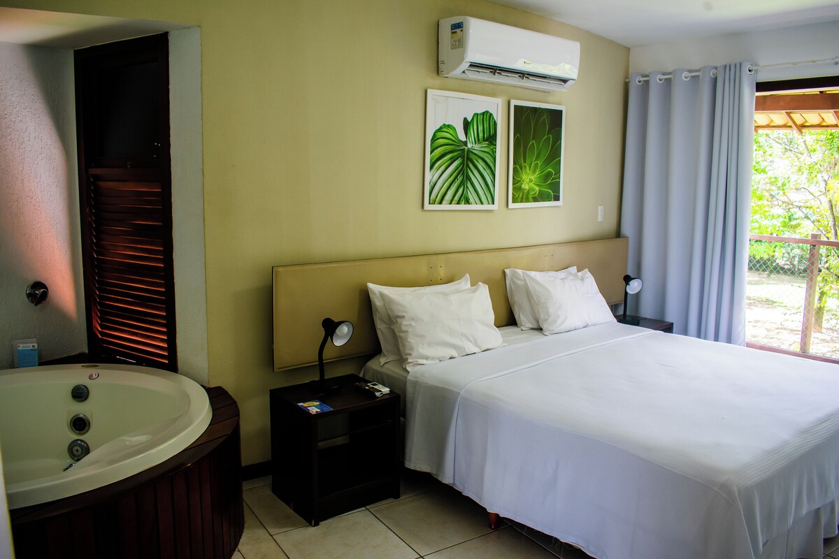 Ein Hotelzimmer mit Bett, Nachttisch, Badezimmer mit Badewanne und Fenster mit Vorhängen.