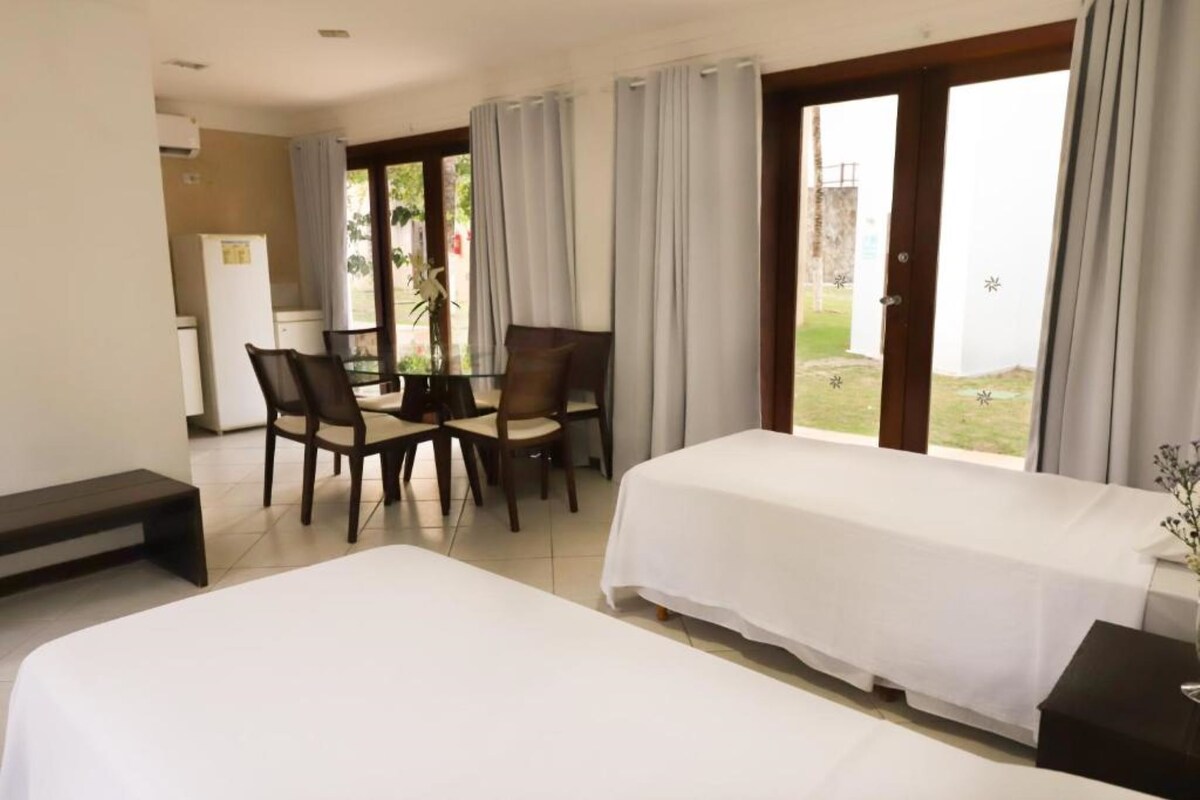 Ein Hotelzimmer mit zwei Betten, einem Esstisch mit Stühlen, einem Kühlschrank und großen Fenstern mit Vorhängen.