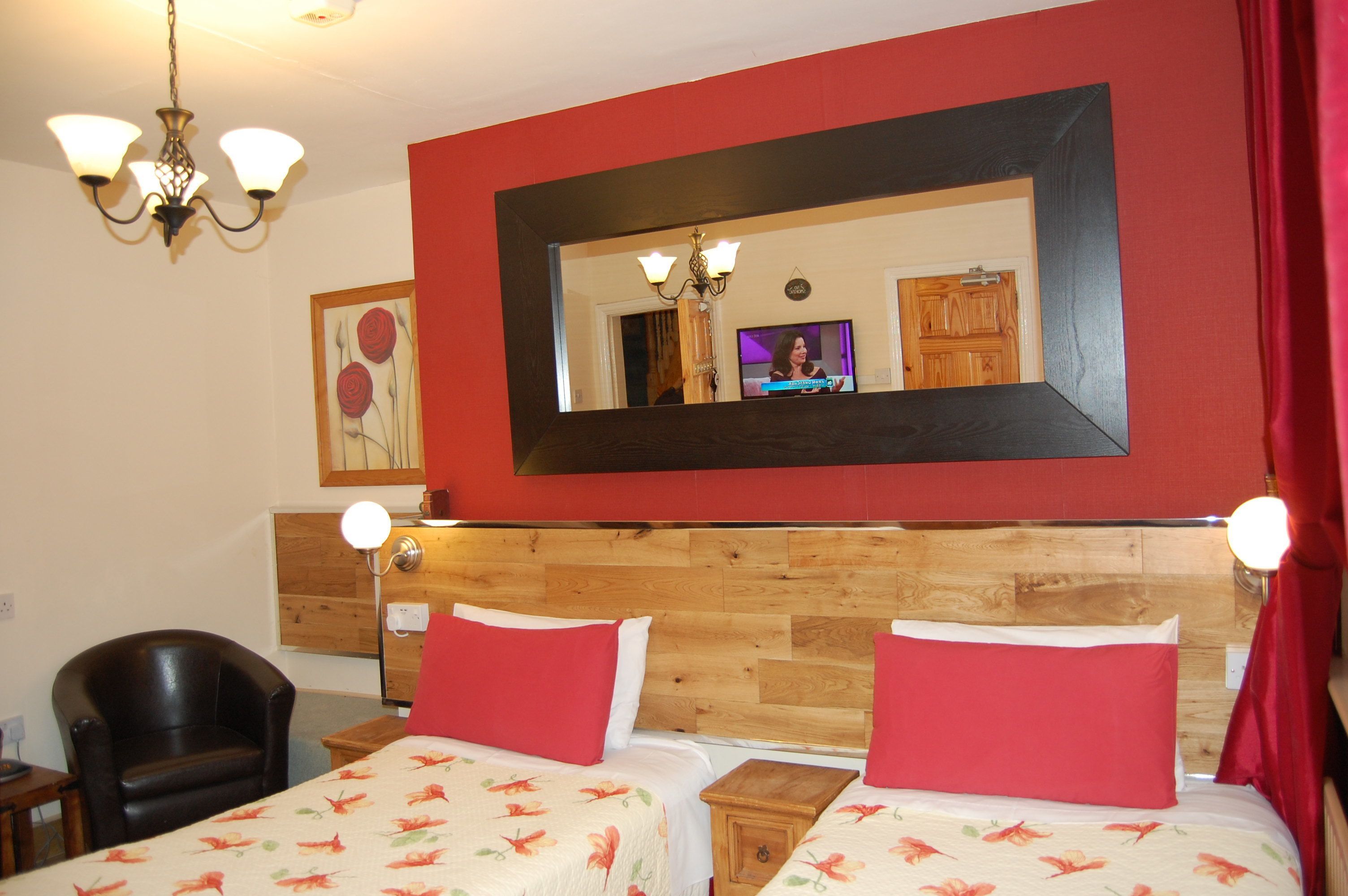 Quarto de hotel com duas camas, cabeceira de madeira, parede vermelha, lustre, televisão e espelho.