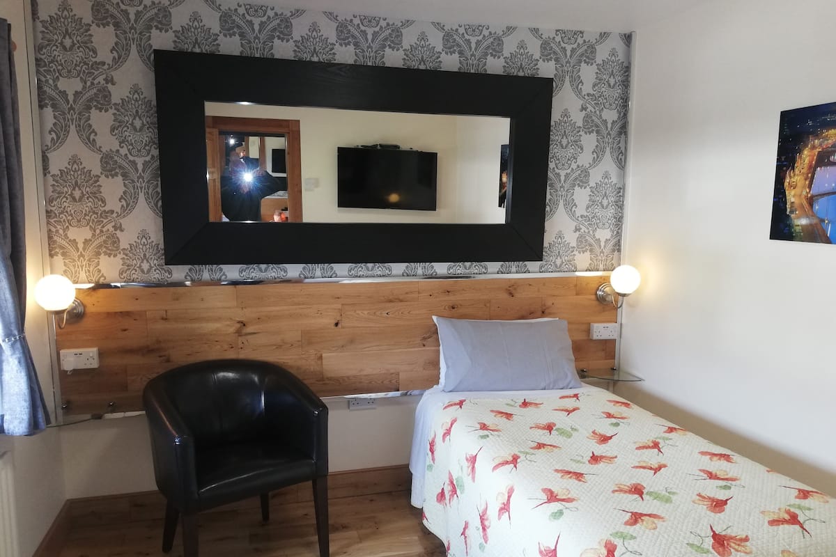 Une chambre à coucher avec une tête de lit en bois, un lit recouvert d’une couvre-lit à motifs floraux, une chaise noire et un grand miroir au-dessus du lit.