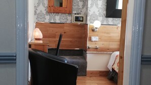 Ein Zimmer mit Holzboden, einem schwarzen Stuhl, einem Holzkasten, einem Spiegel, einem Heizkörper und einem Bett mit weißer Decke.