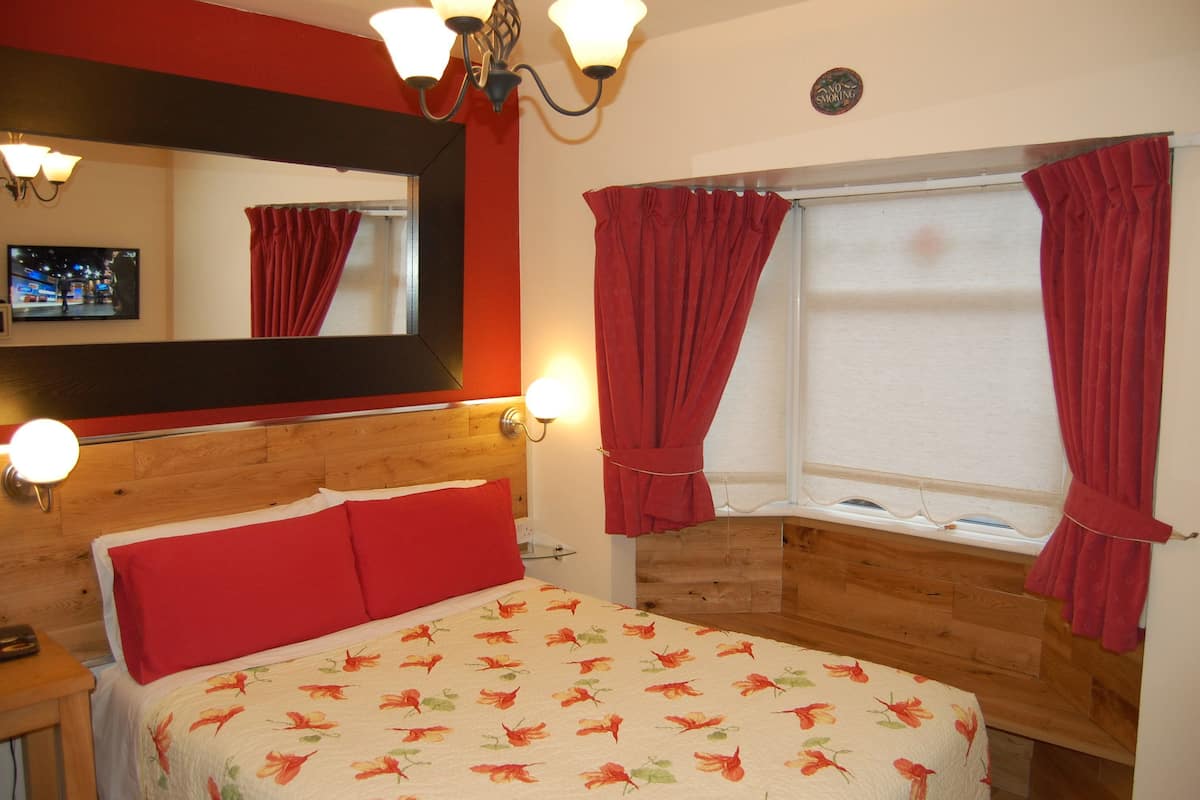 Une chambre à coucher avec une tête de lit en bois, un lit avec des oreillers rouges, une couvre-lit à motifs floraux, un miroir, un téléviseur fixé au mur et des rideaux rouges aux fenêtres.