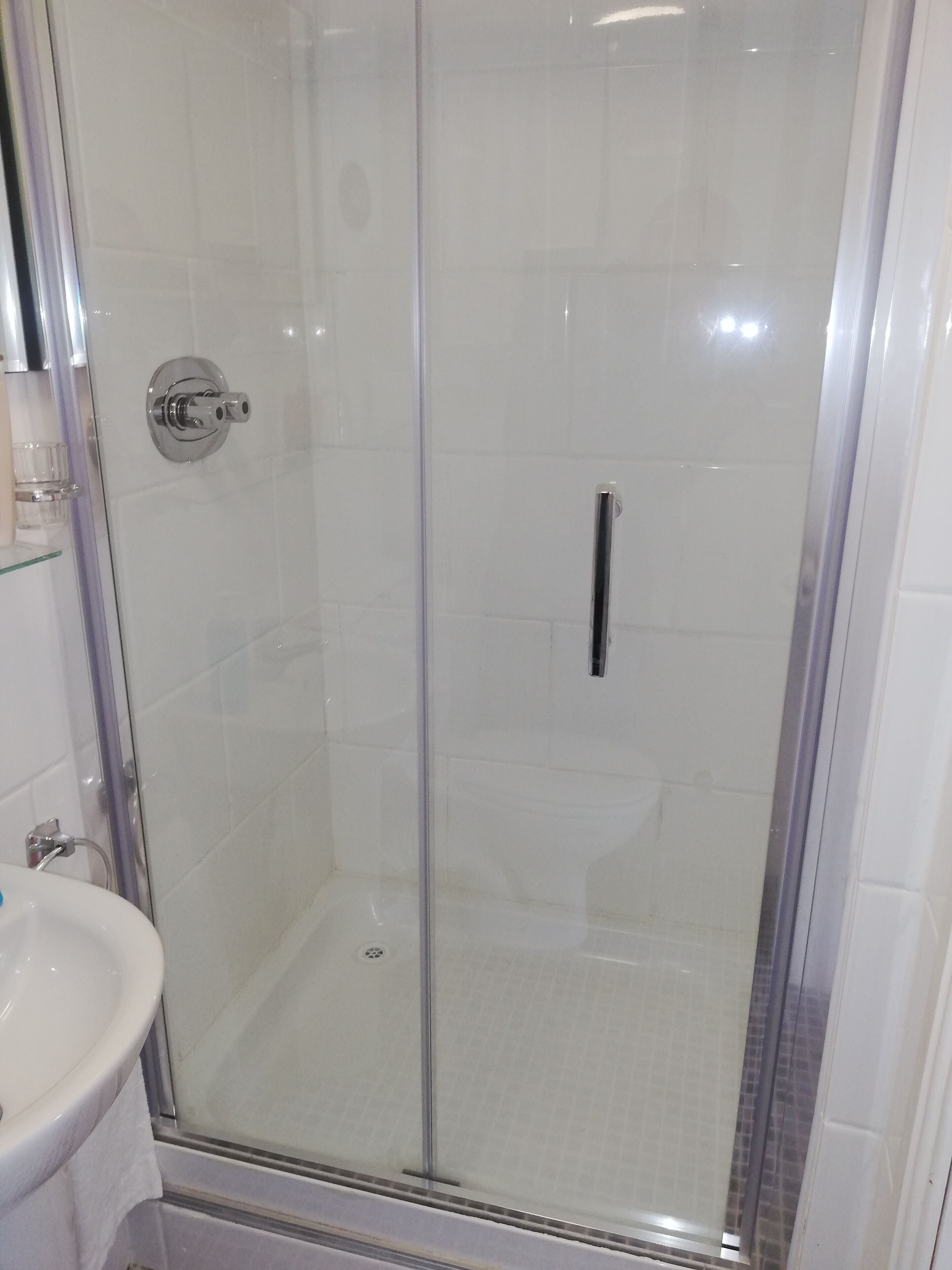 Eine begehbare Dusche mit Glaswand, einem Chrom-Armaturenknopf, einem weißen Waschbecken und einer gefliesten Wand.