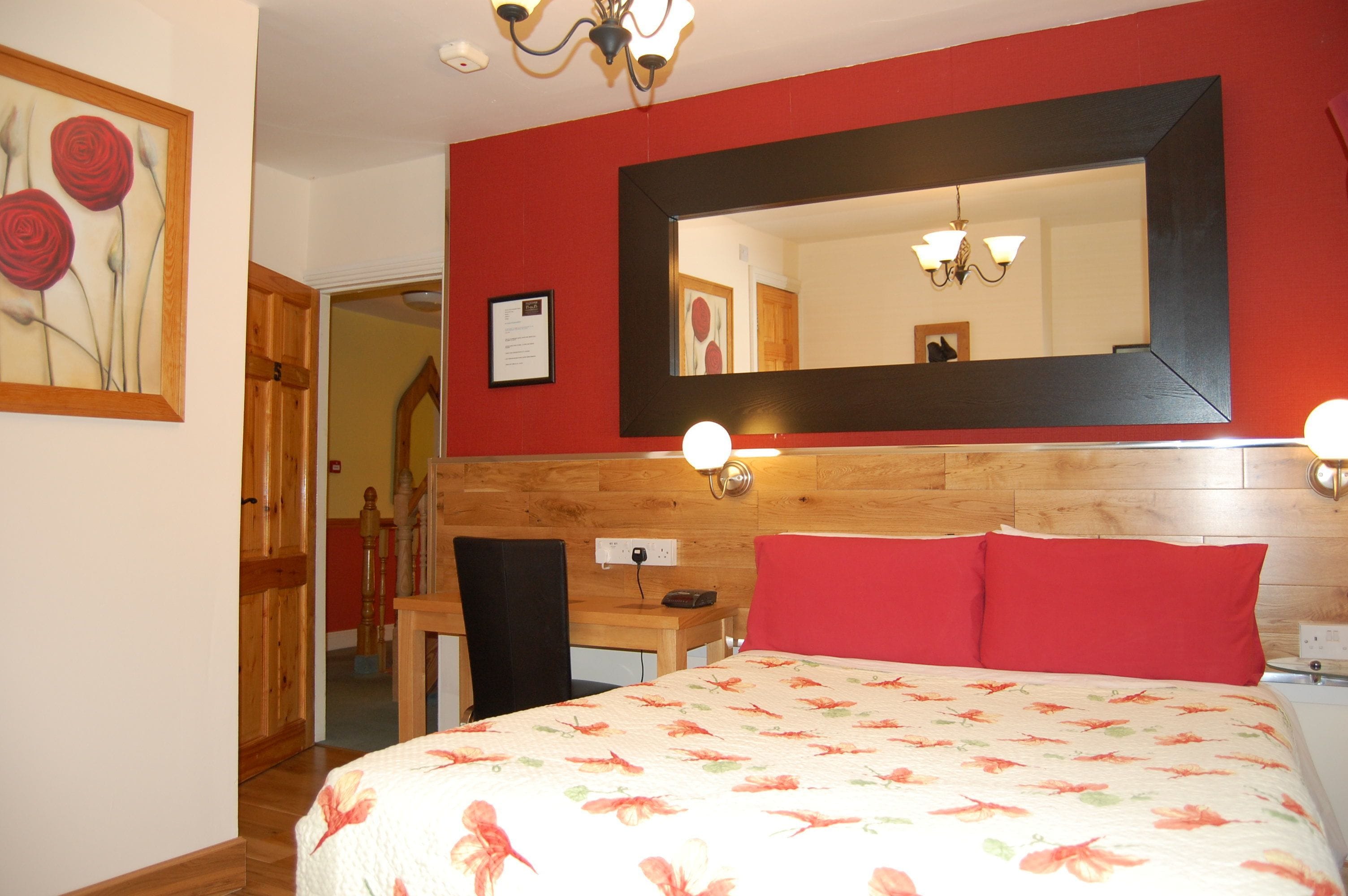 A bedroom with a large bed, a wooden desk, a chair, a mirror, and a red wall.