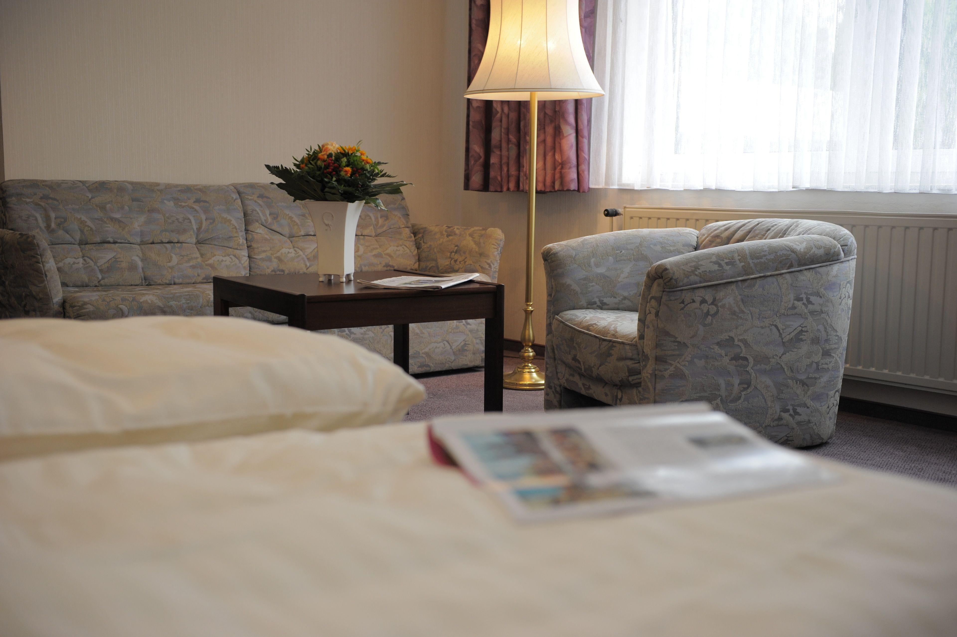 A hotel room with a bed, a sofa, a chair, a table with a vase of flowers, and a lamp.