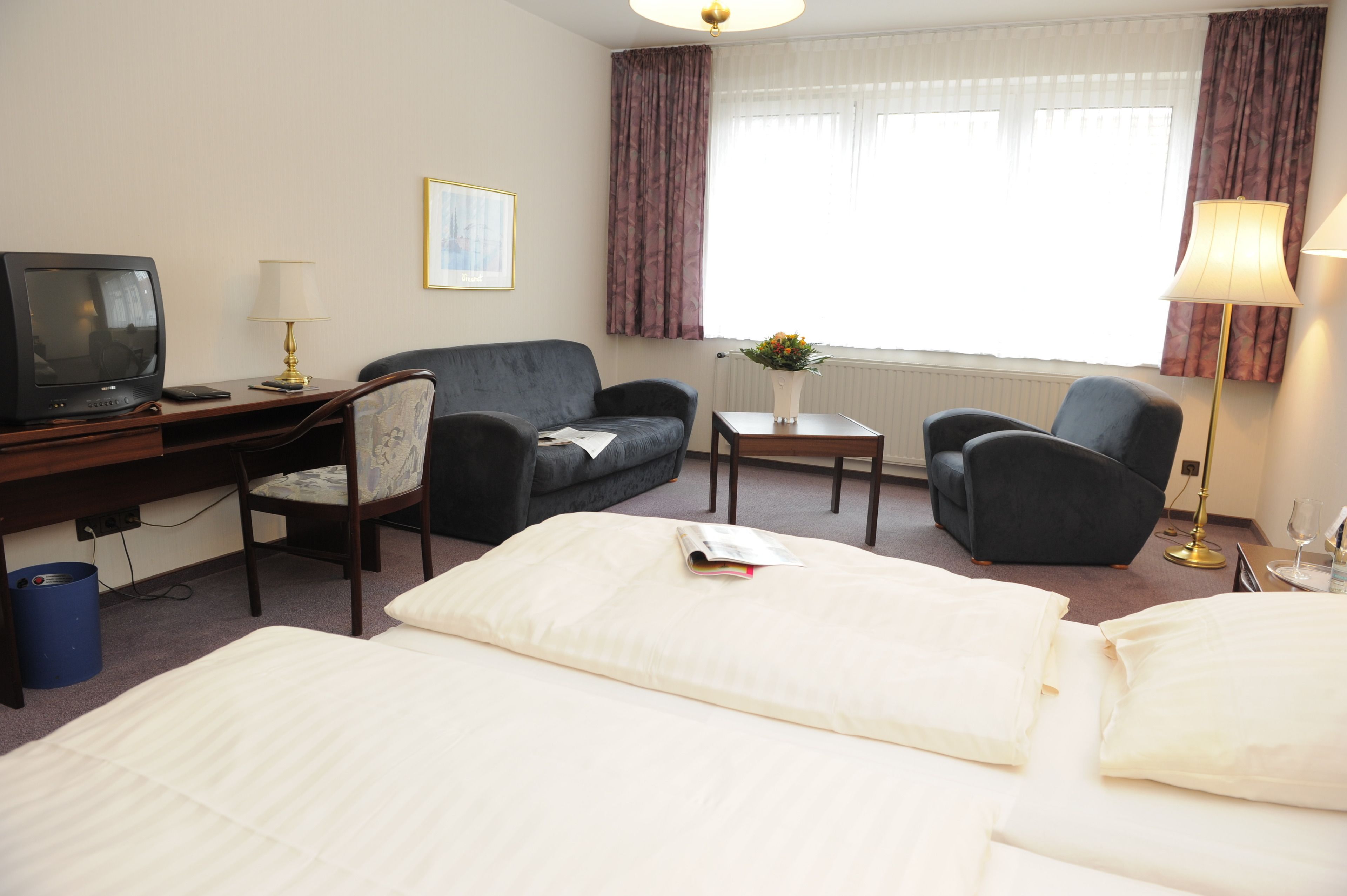 Habitación de hotel con cama, dos sillones, escritorio con televisor, lámpara y ventana con cortinas.