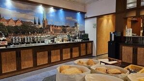 Un vestíbulo de hotel con un gran mural de la ciudad, una cafetería con diferentes máquinas y recipientes de café, y zonas de estar con cestas de pan.