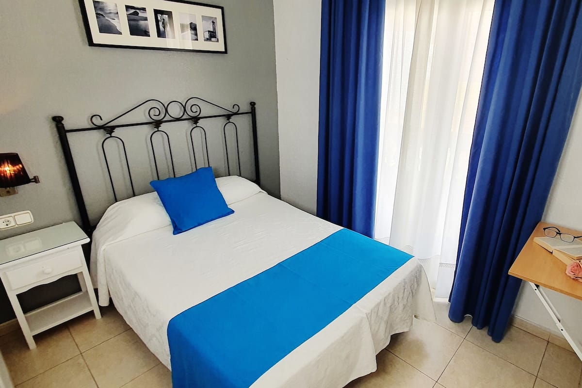 A neatly made bed with a blue accent pillow, a bedside table with a lamp, a small wooden side table with a book and a rose, and a TV mounted on the wall.